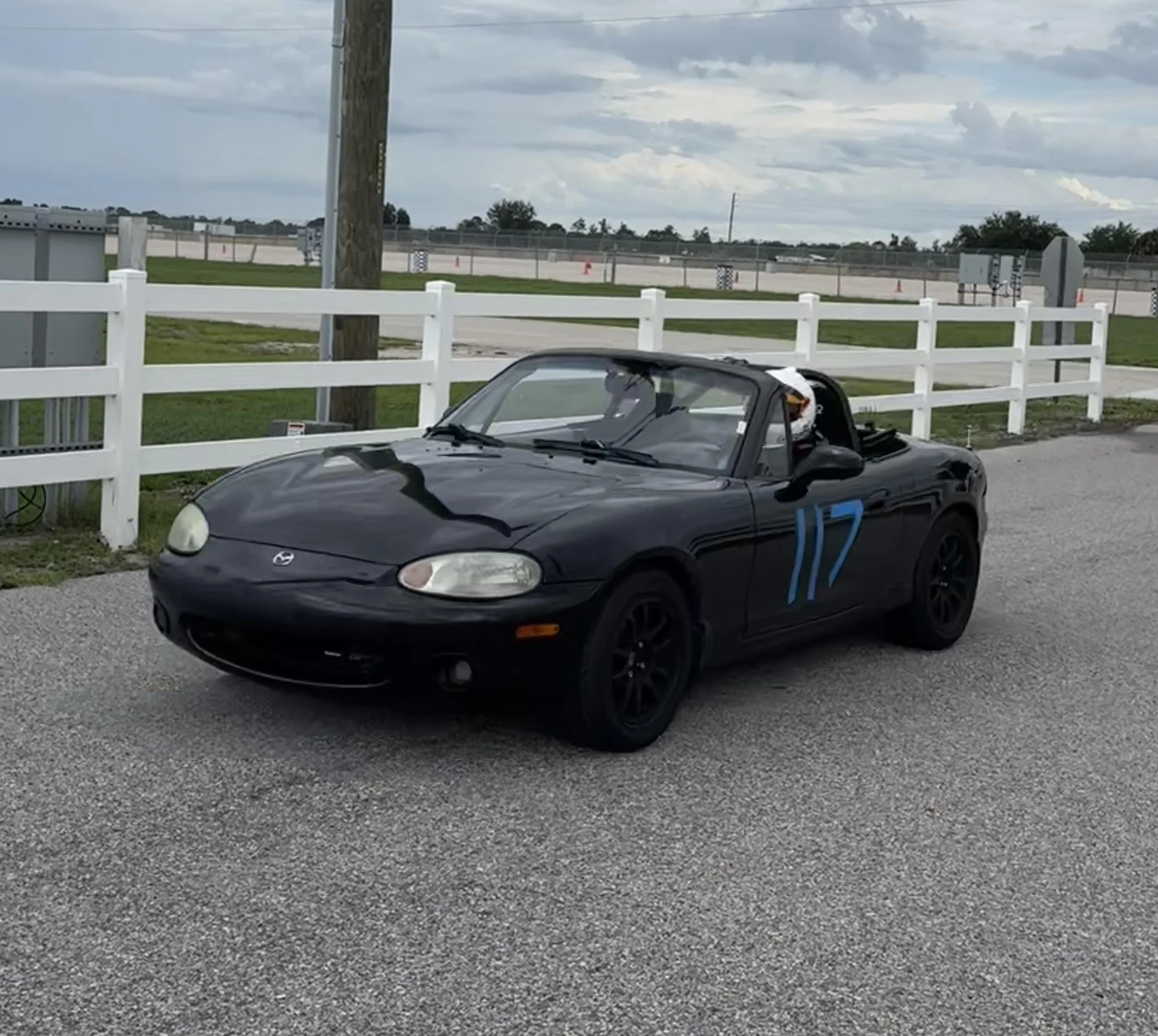 A black Mazda convertible car parked on a gravel surface with a white fence behind it and electrical meters mounted on the fence. In the background, there is an open area with a chain-link fence and orange traffic cones, under a cloudy sky.