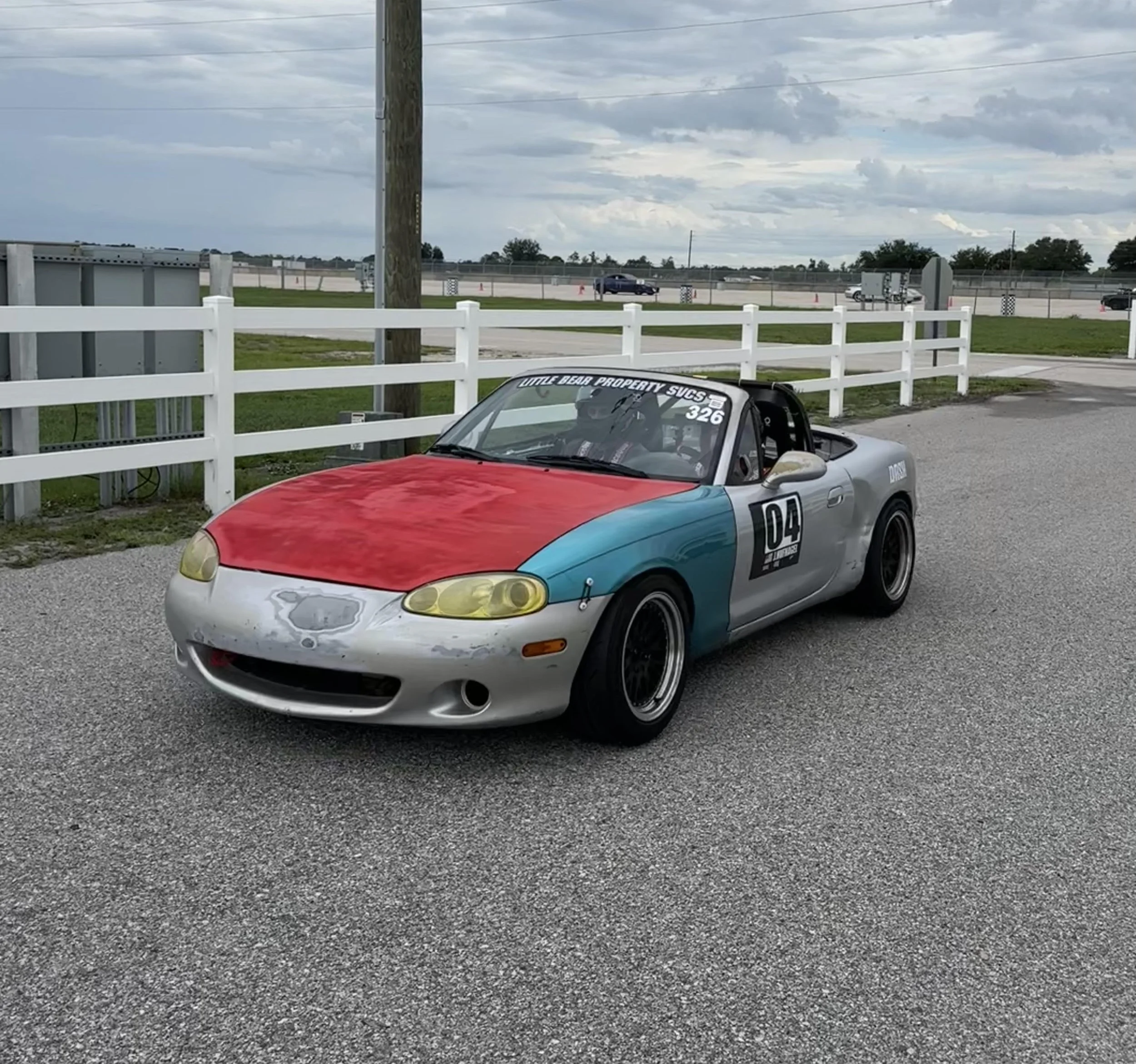 A colorful sports car with a red hood, yellow headlights, and a partially visible number 04 on the side, parked in front of a white fence and utility pole at a race track under a cloudy sky.