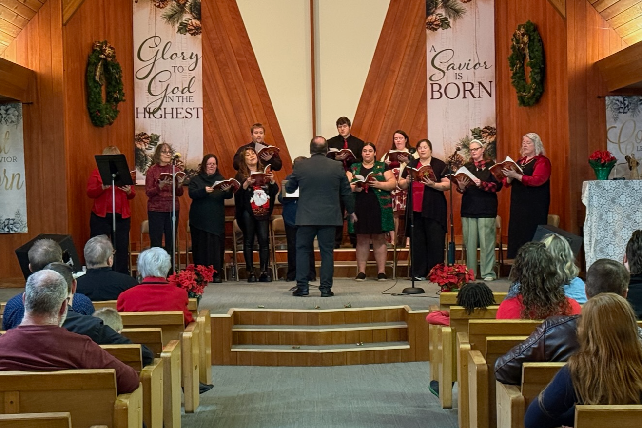 A choir standing on stage in Christmas attire, led by a conductor, performing in a church decorated for Christmas with wreaths, poinsettias, and banners that read 'Glory to God in the Highest' and 'A Savior is Born.'