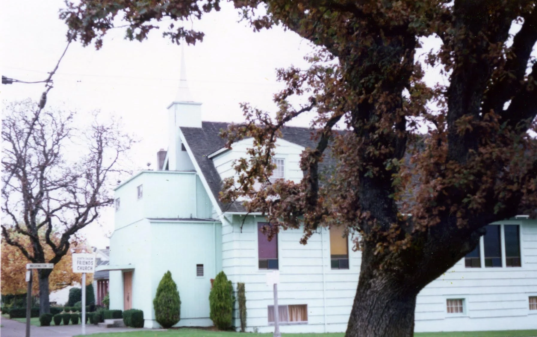 A white church building with a steeple and cross visible above the roof, surrounded by trees with brown autumn leaves, and a street sign that reads "FRIENDS CHURCH".