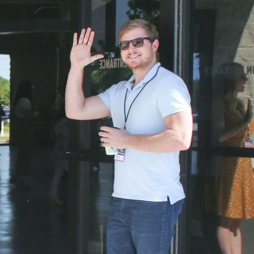 A young man with sunglasses, a white polo shirt, and jeans, smiling and waving while standing outside a building.