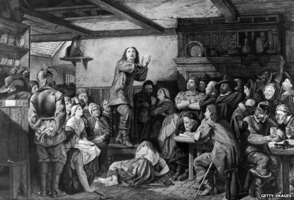 A black and white illustration of a lively tavern or pub scene with a man standing on a table gesturing passionately, surrounded by a diverse group of men, women, and children listening and watching intently. The room has shelves with bottles and a fireplace, suggesting a rustic, historic setting.