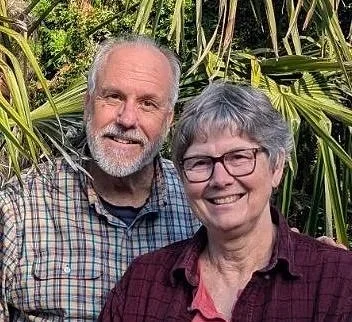 A smiling man and woman standing outdoors with greenery in the background.