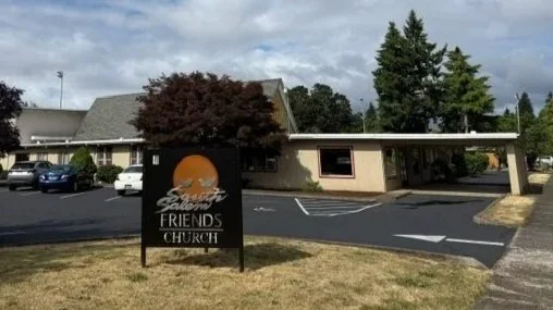 Sign for Shoreline Friends Church in a parking lot with parked cars, a building, and trees in the background.
