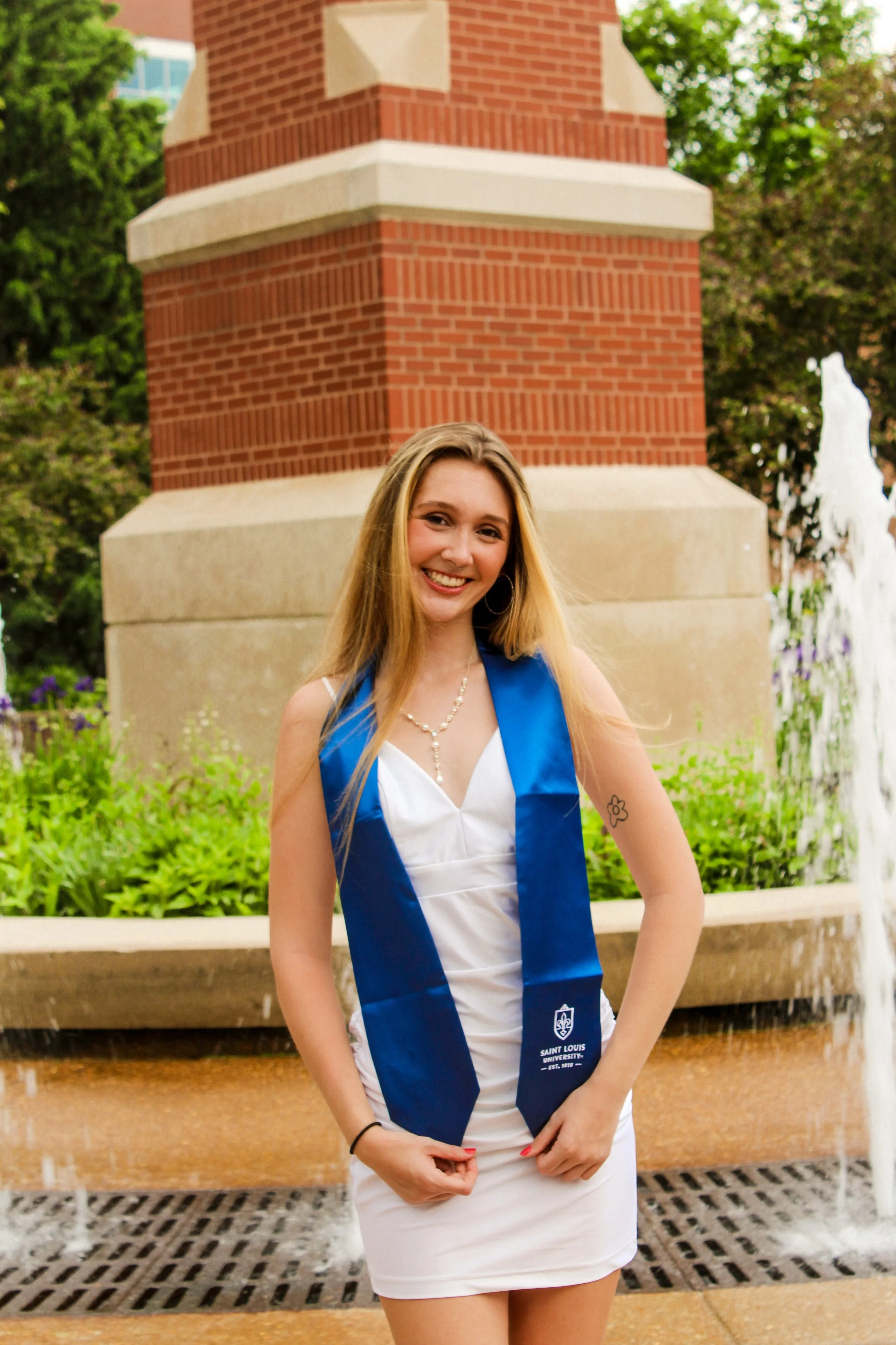 A young woman with blonde hair smiling at the camera, wearing a white dress and a blue graduation stole with the Saint Louis University logo, standing in front of a brick fountain and greenery.
