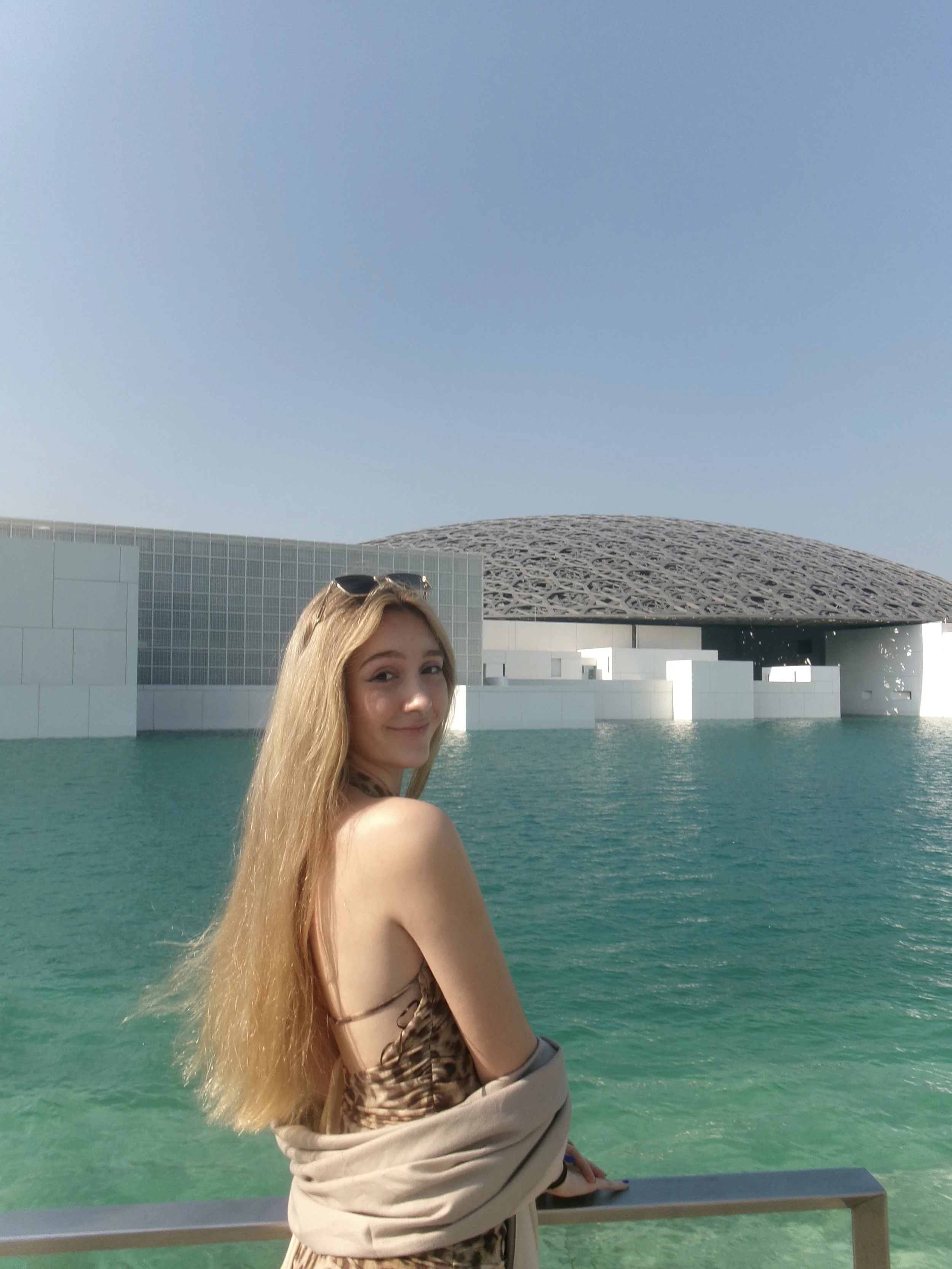 A young woman with long blonde hair sitting on a railing by a body of water, with a modern architectural building and a domed structure in the background on a sunny day.