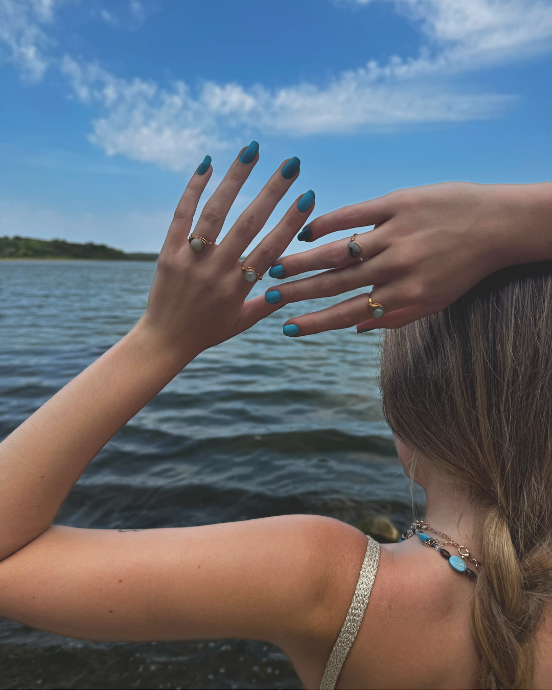 Two women with painted nails and jewelry, holding hands near a body of water under a blue sky with clouds.