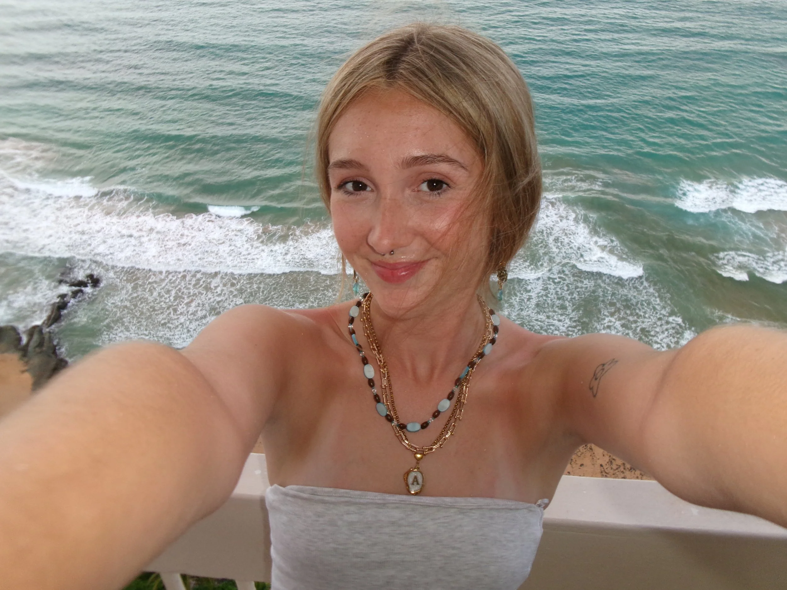 A woman taking a selfie at the beach during daytime with ocean waves in the background. She has shoulder-length blonde hair, is smiling, and wearing multiple necklaces and earrings.
