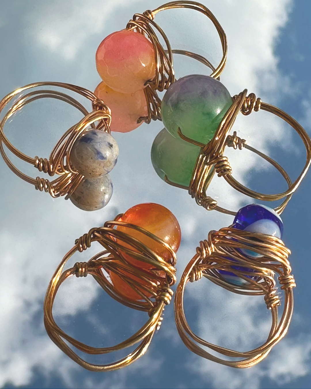Colorful beads wrapped in gold wire jewelry held up against a cloudy blue sky.