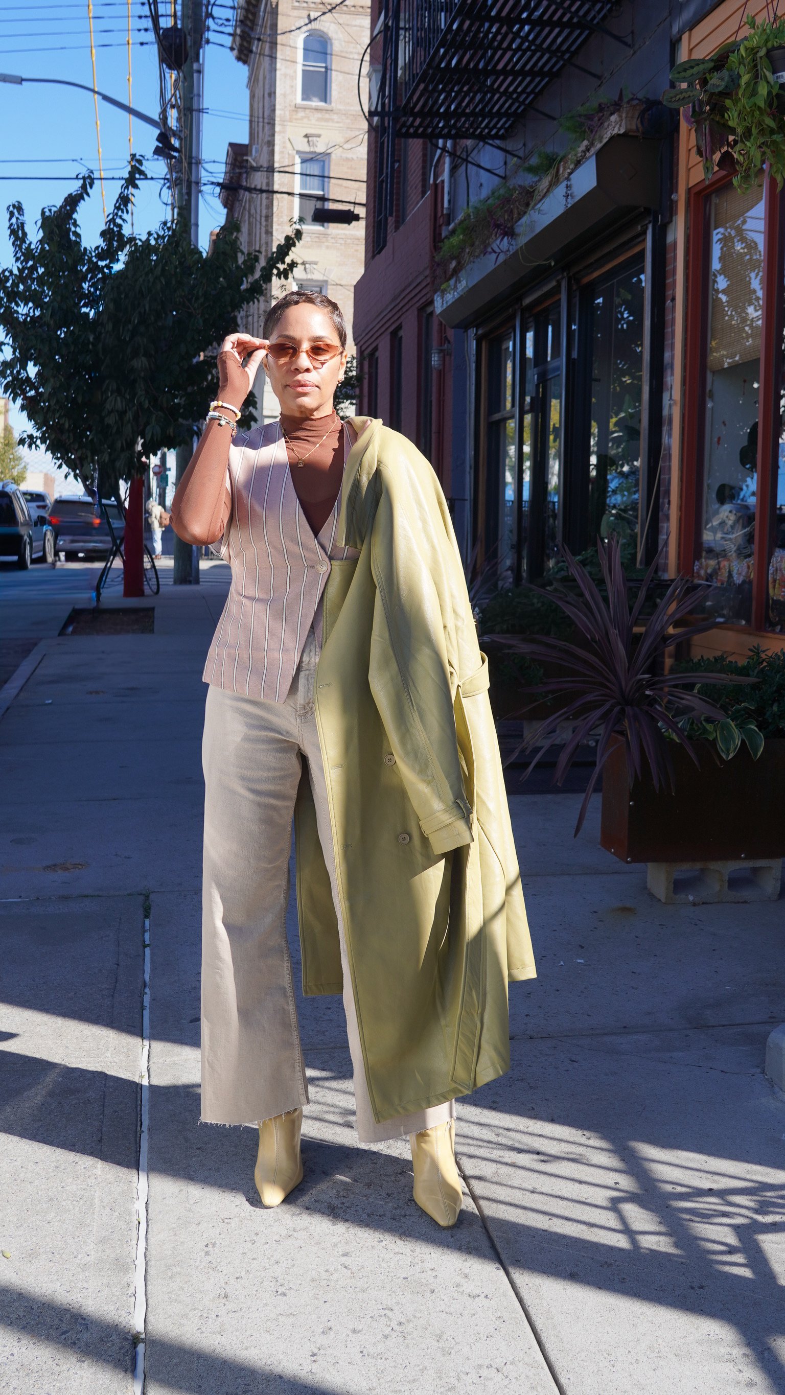 Woman standing on sidewalk wearing beige pants, a brown top, a striped vest, yellow coat draped over her shoulder, yellow sunglasses, jewelry, and beige boots, with buildings, trees, and parked cars in the background.