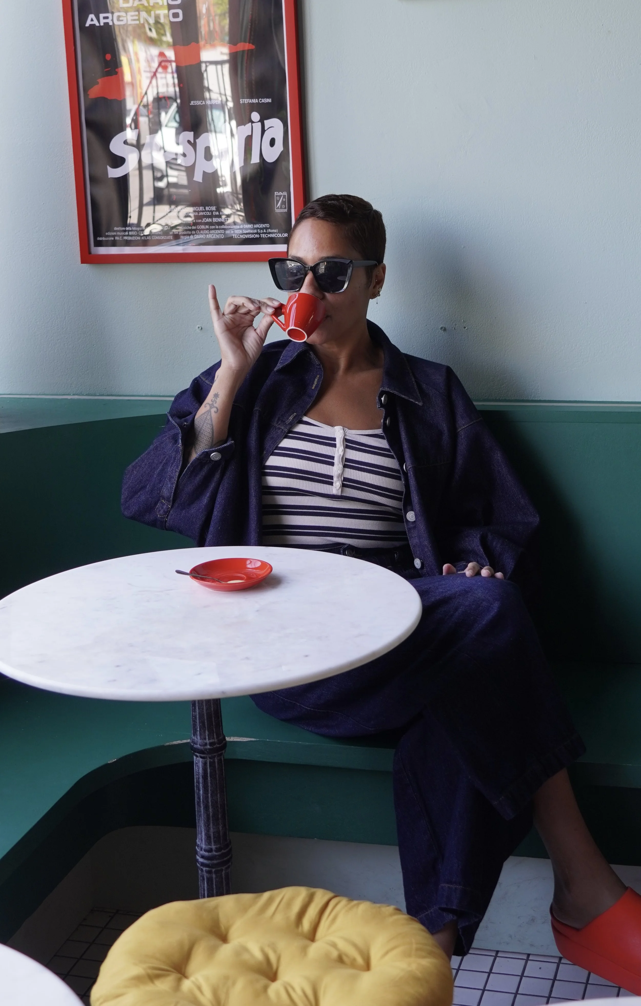 Person sitting at a table in a cafe, wearing sunglasses, drinking from a small red cup, dressed in a striped shirt and dark jacket, with a tattoo on her left arm. There is a vibrant yellow cushion on the seat near her and a framed poster on the wall behind her.