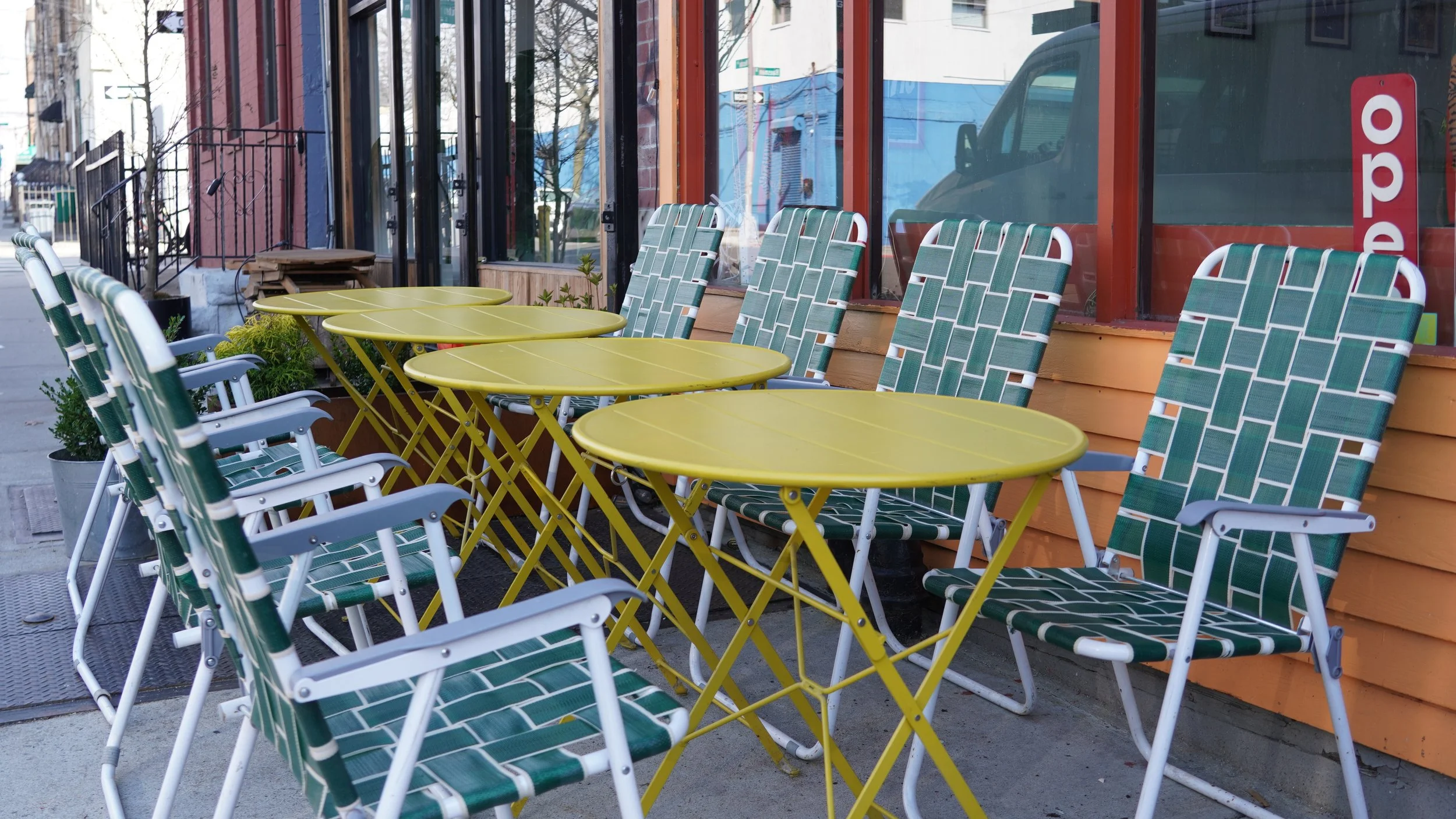 Outdoor seating area with yellow round tables and green patterned chairs outside a restaurant or cafe, with large windows reflecting street scenery.