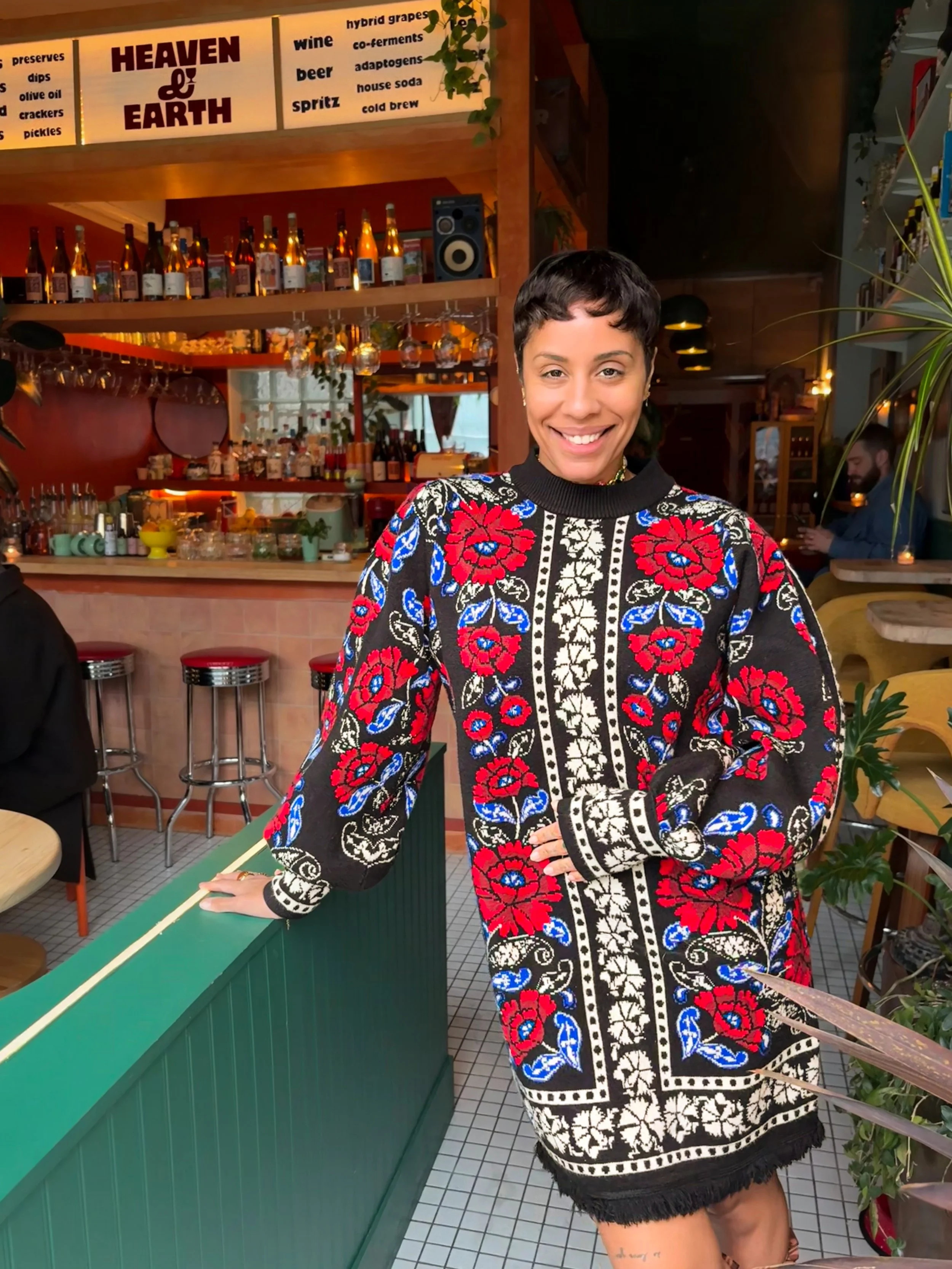 Smiling woman with short black hair wearing a black, red, and blue floral patterned dress, standing in a restaurant or bar with a green tiled counter and shelves of bottles and glasses behind her.