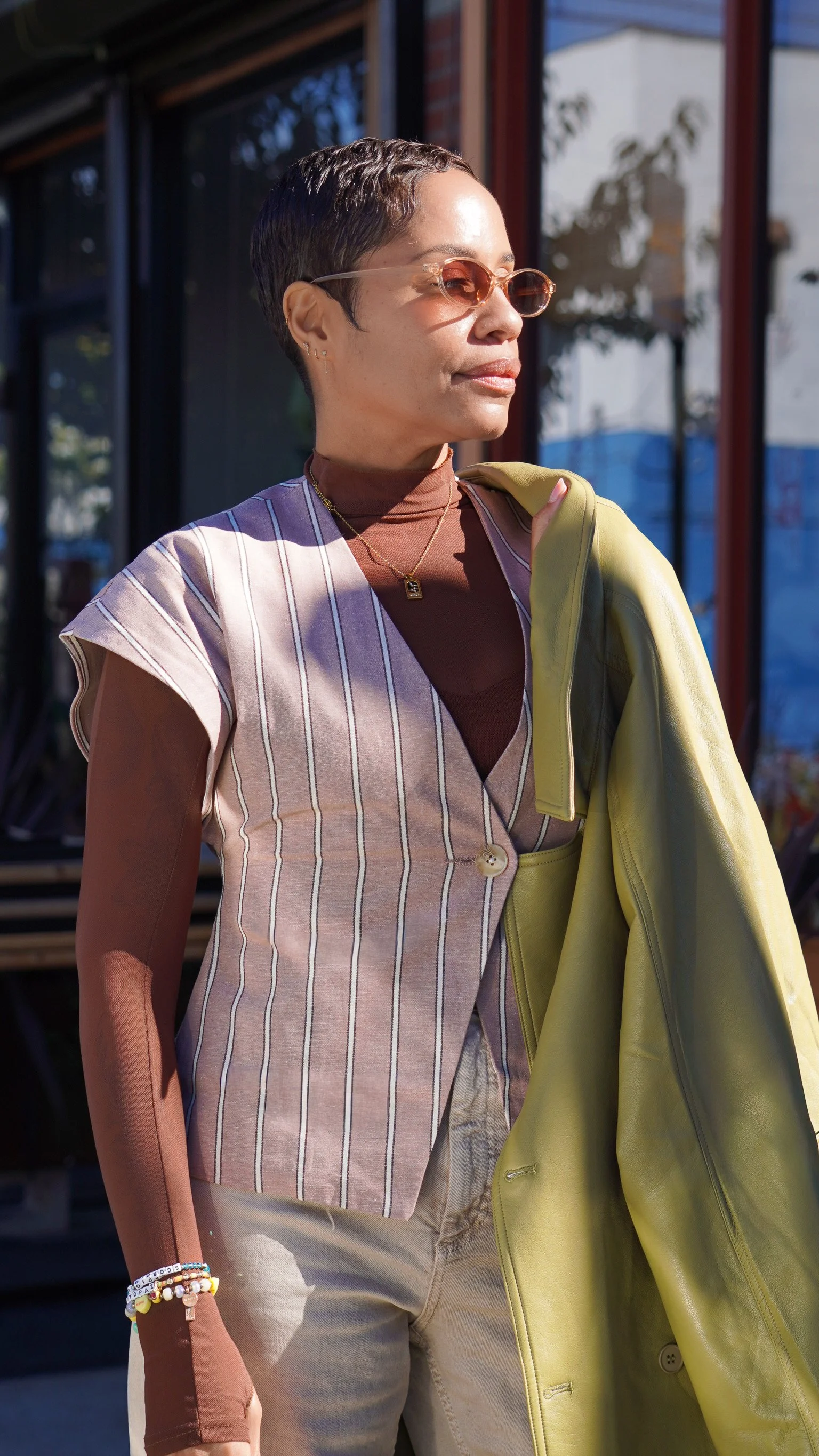 Woman with short dark hair wearing sunglasses, a striped vest, jewelry, and carrying a yellow jacket outside.