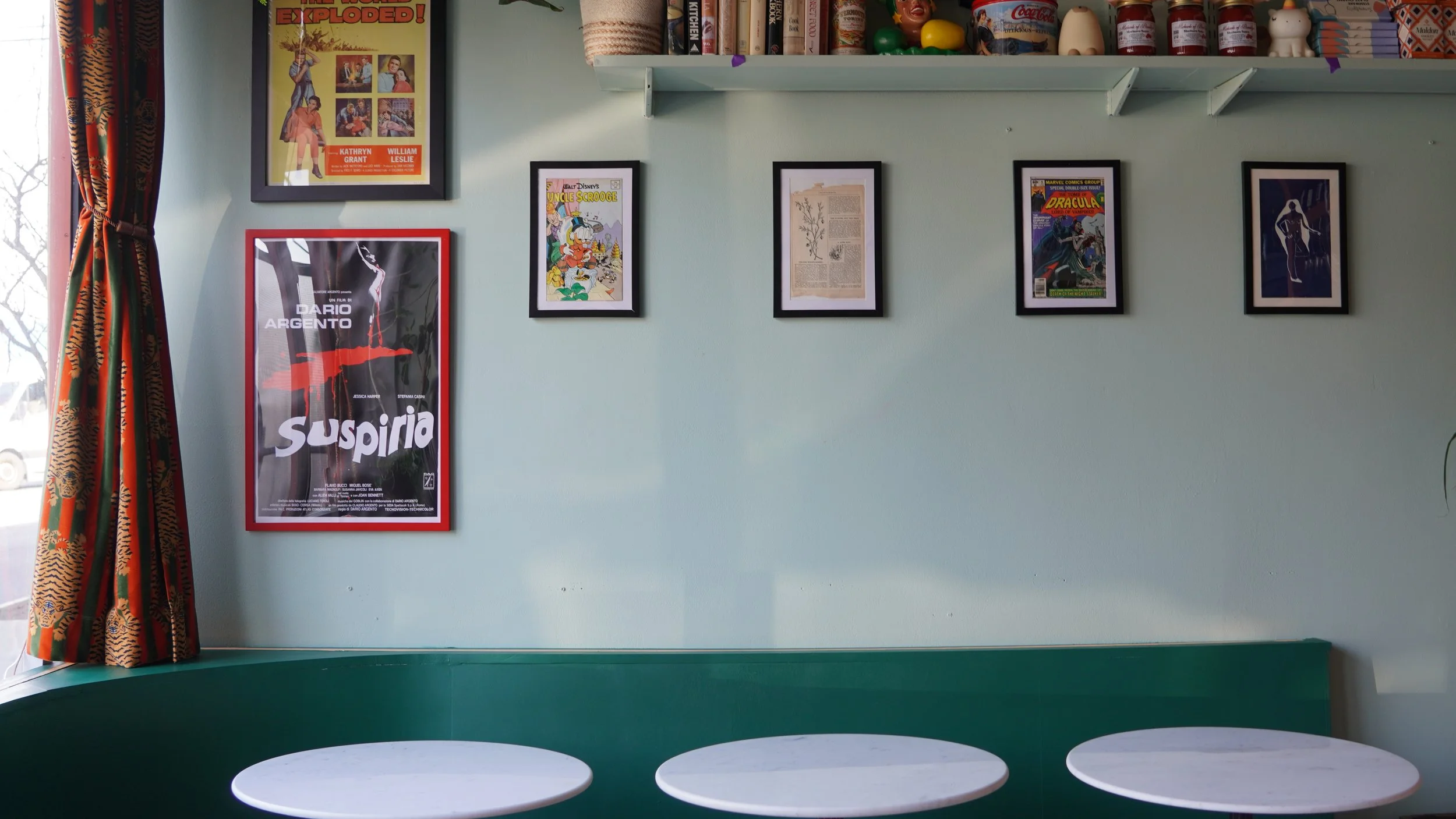 A wall with framed posters and artwork, including a 'Suspiria' movie poster, a 'Disney's Uncle Scrooge' comic, and other framed pieces. A bookshelf with books and toys is at the top, with a window partially covered by patterned curtains on the left. The bottom features a green bench with four white round tables in front.