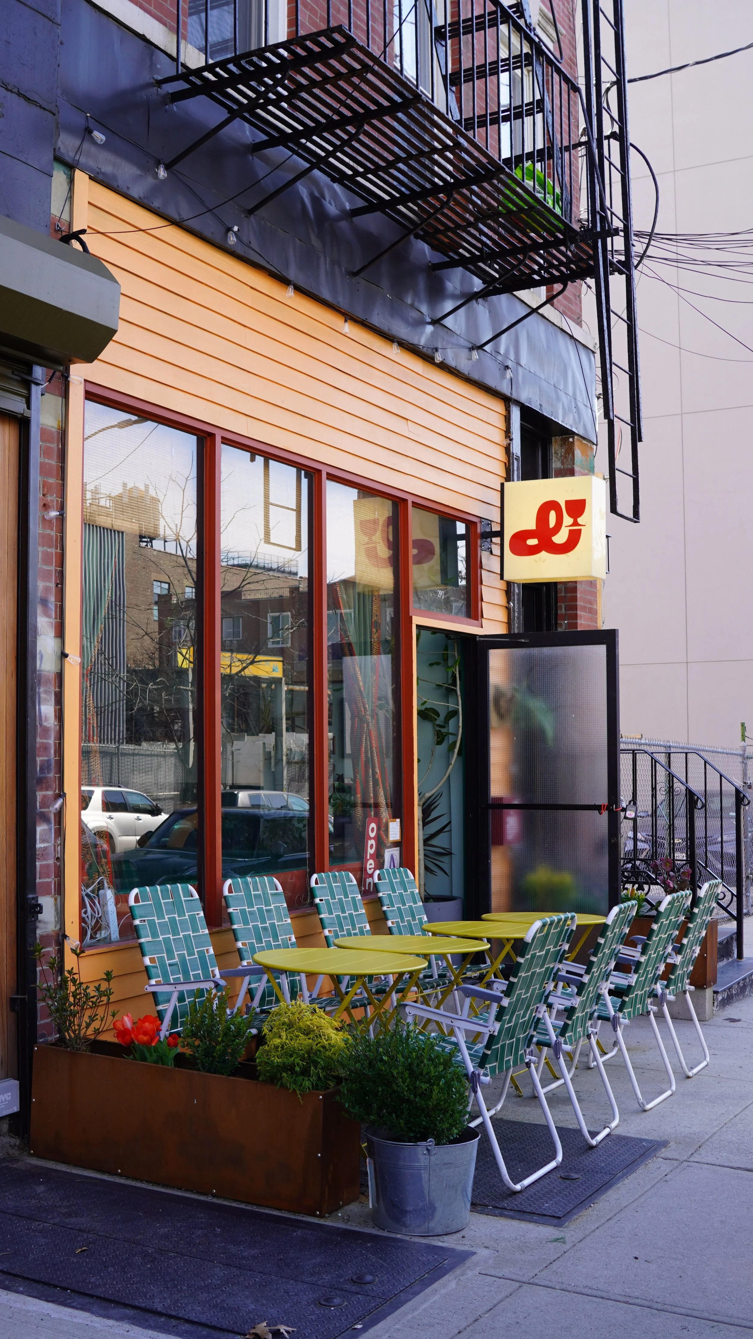 Outdoor patio seating area with green metal tables and chairs with striped cushions in front of a restaurant with large glass windows and an orange facade. A planter with yellow and red flowers and green plants is near the seating. The restaurant has a sign with a red logo, a black door, and a small window.