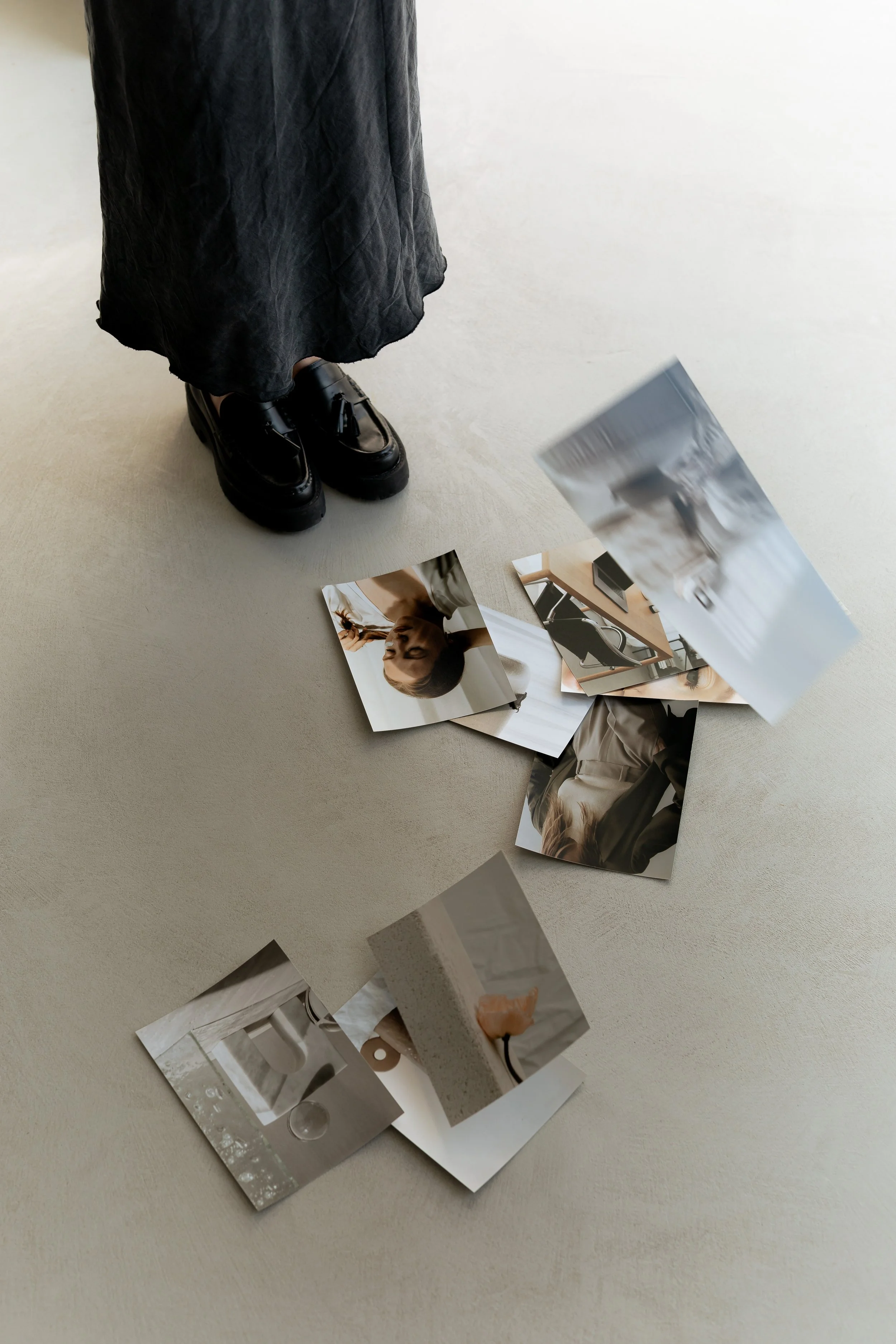 A person standing on a beige carpet wearing a long black skirt and shiny black platform shoes, with scattered aesthetic photographs on the floor.