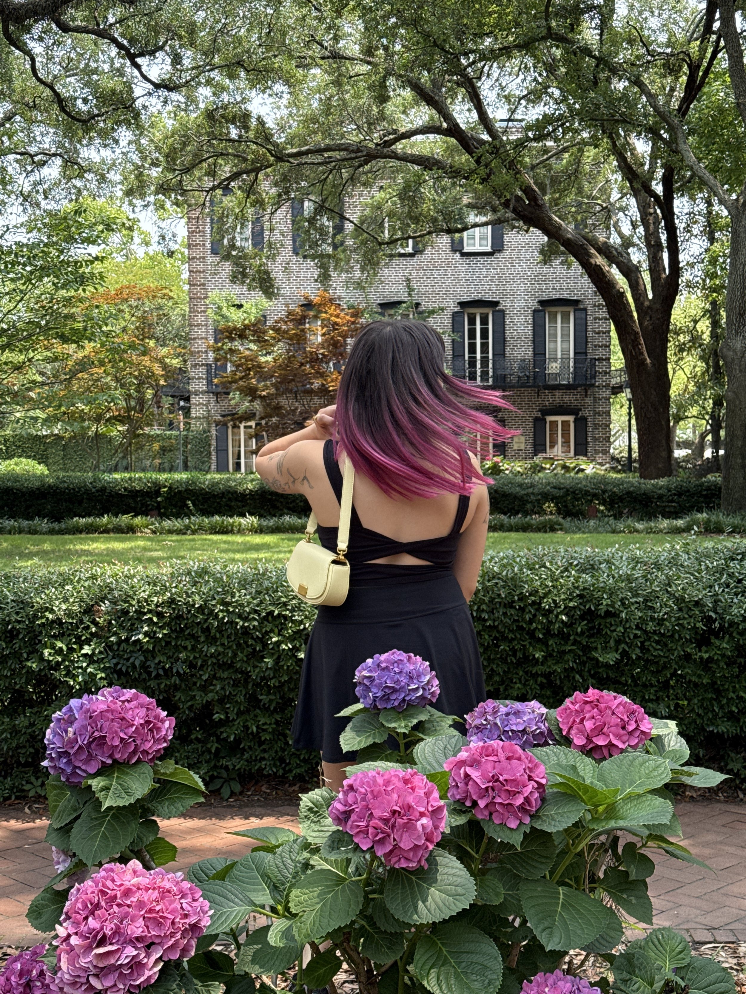A woman with brown hair with pink ends, wearing a black dress and a yellow purse on her shoulder, stands in a garden amidst pink and purple hydrangea flowers. She faces away from the camera, with trees and a large brick house in the background.