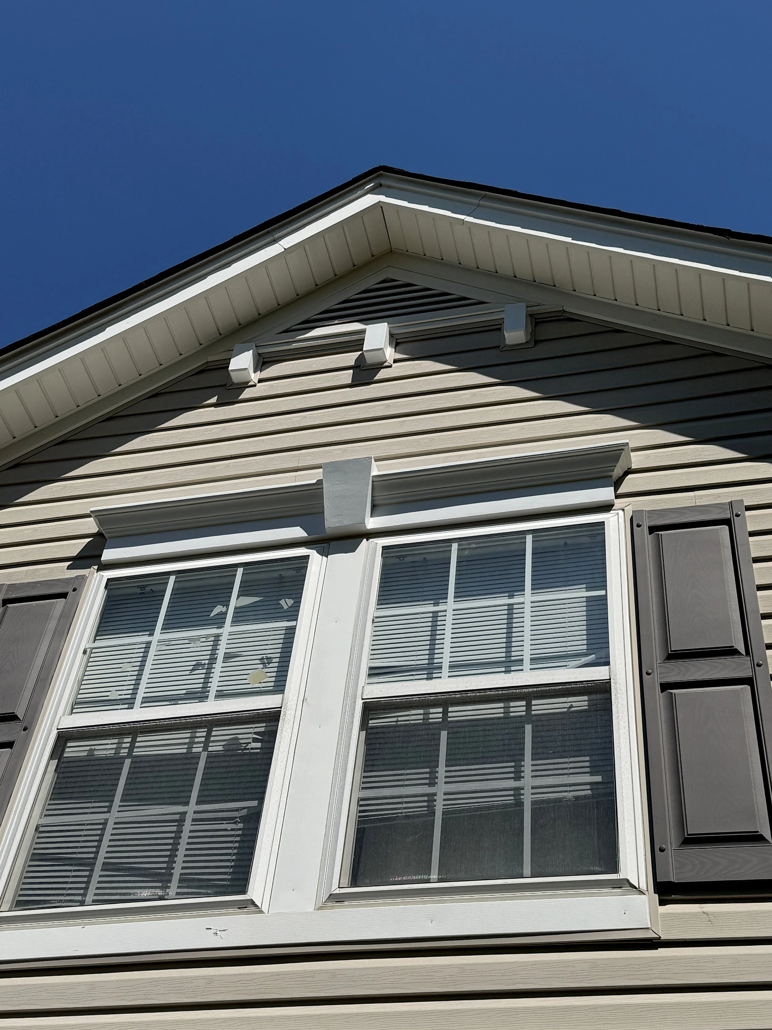 Close-up of beige house siding with a large window and shutters, under a clear blue sky, with the roof and part of the eaves visible.