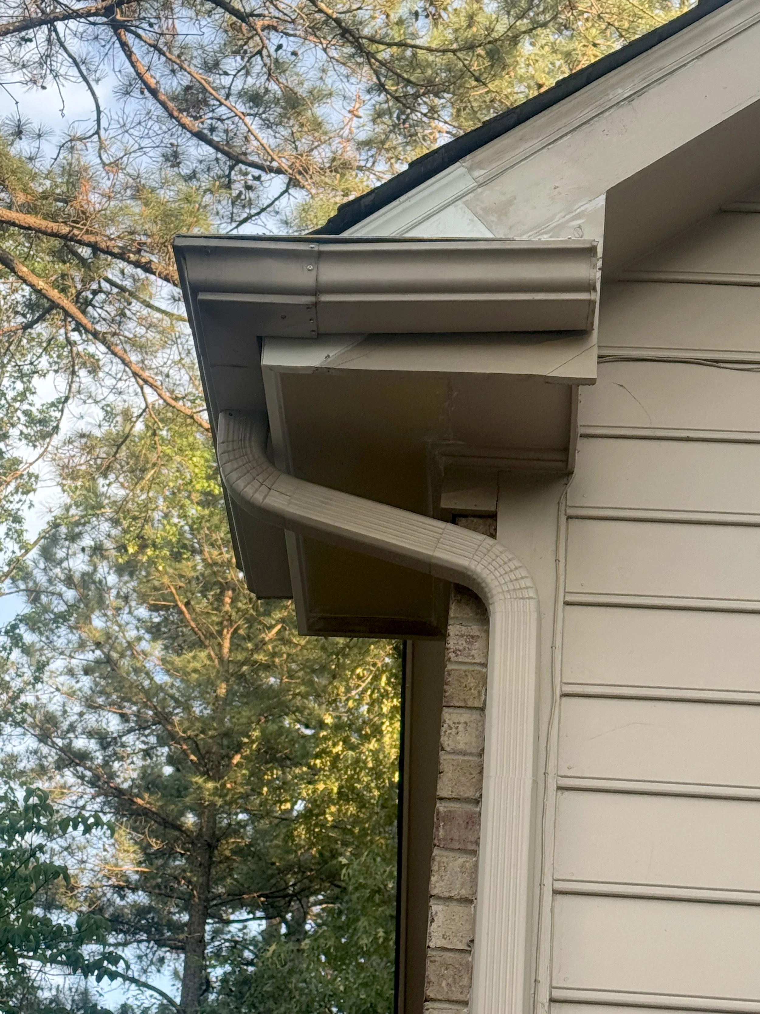 View of a house's roofline with a vinyl gutter, soffit, and siding. Trees with green leaves and branches are in the background, and the sky is visible.