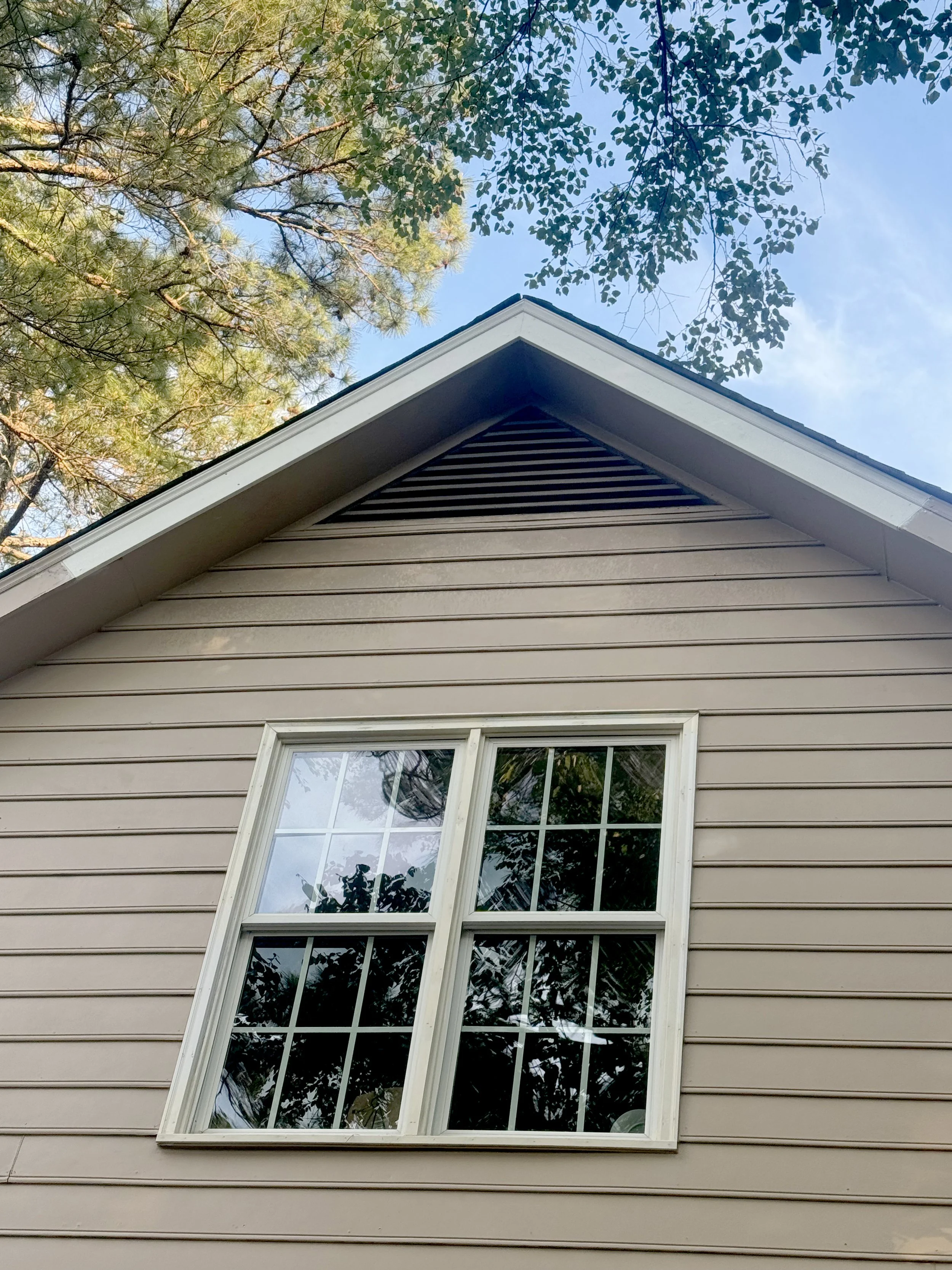 Looking up at the corner of a house with beige siding and white trim, a double-hung window with a white frame, and a triangular attic vent, with green trees and blue sky overhead.