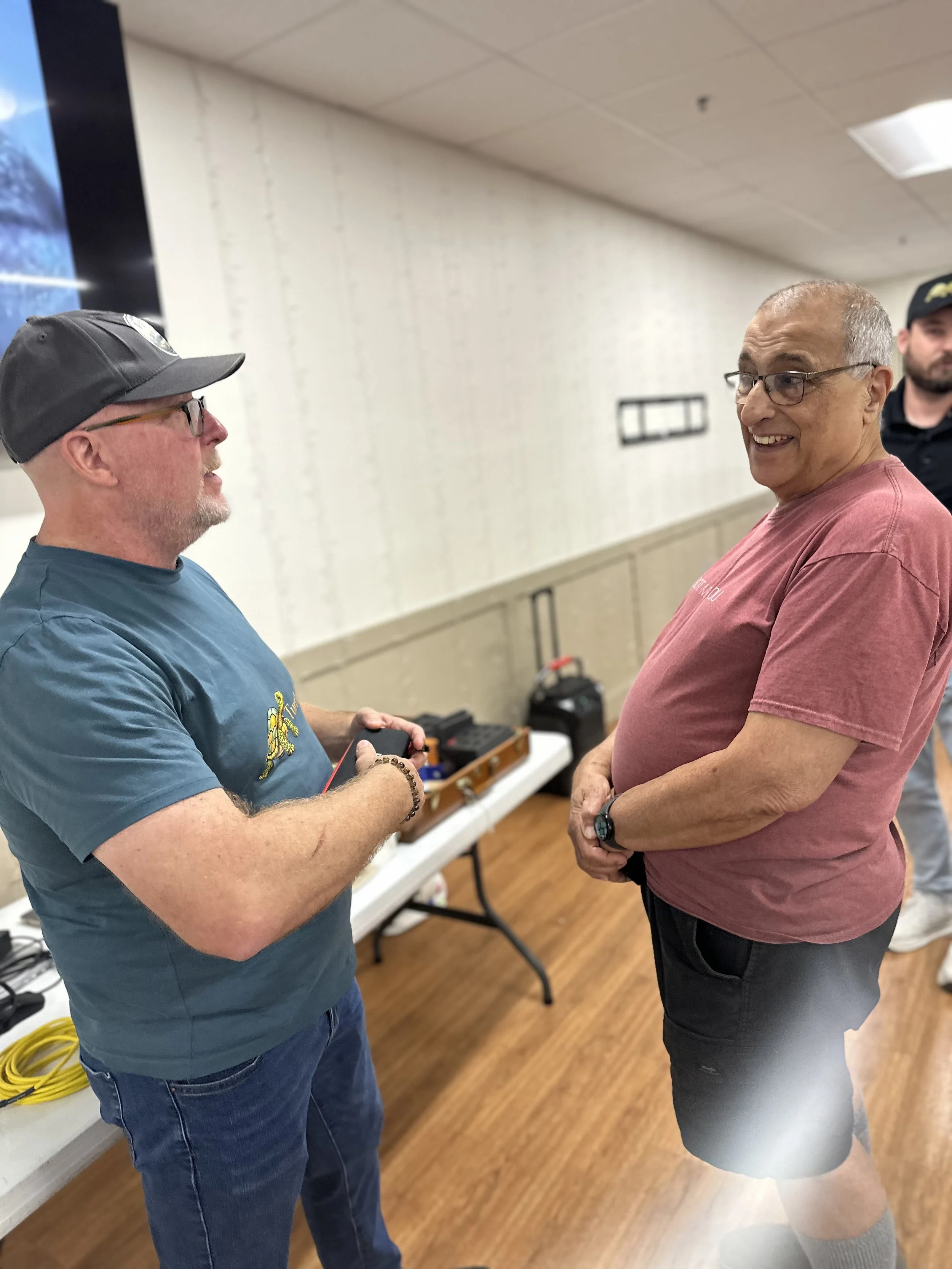 Two men are talking, one holding an object, in a room with a white wall, cupboard, and yellow cable on the floor.