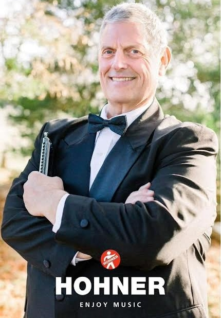 Man in tuxedo holding a large chromatic harmonica outdoors, smiling, with a Hohner promotional logo and text at the bottom.