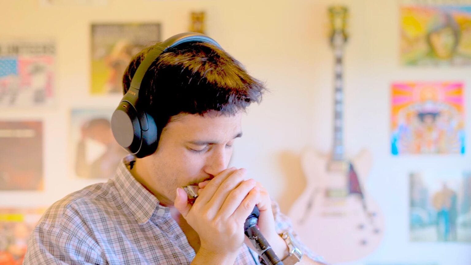 Young man wearing headphones, playing harmonica, with colorful posters and a guitar on the wall behind him.