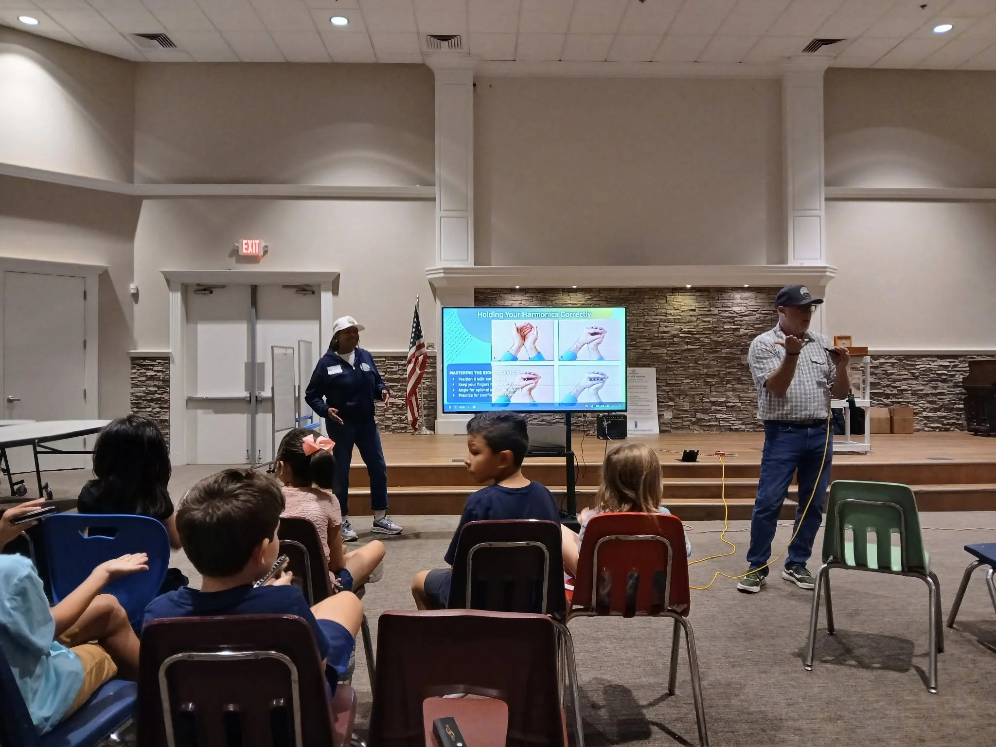 Children seated in chairs watching a presentation in a room with a large screen displaying instructions on how to hold a harmonica correctly. Two instructors, a man and a woman, are standing near the screen, explaining the instructions.
