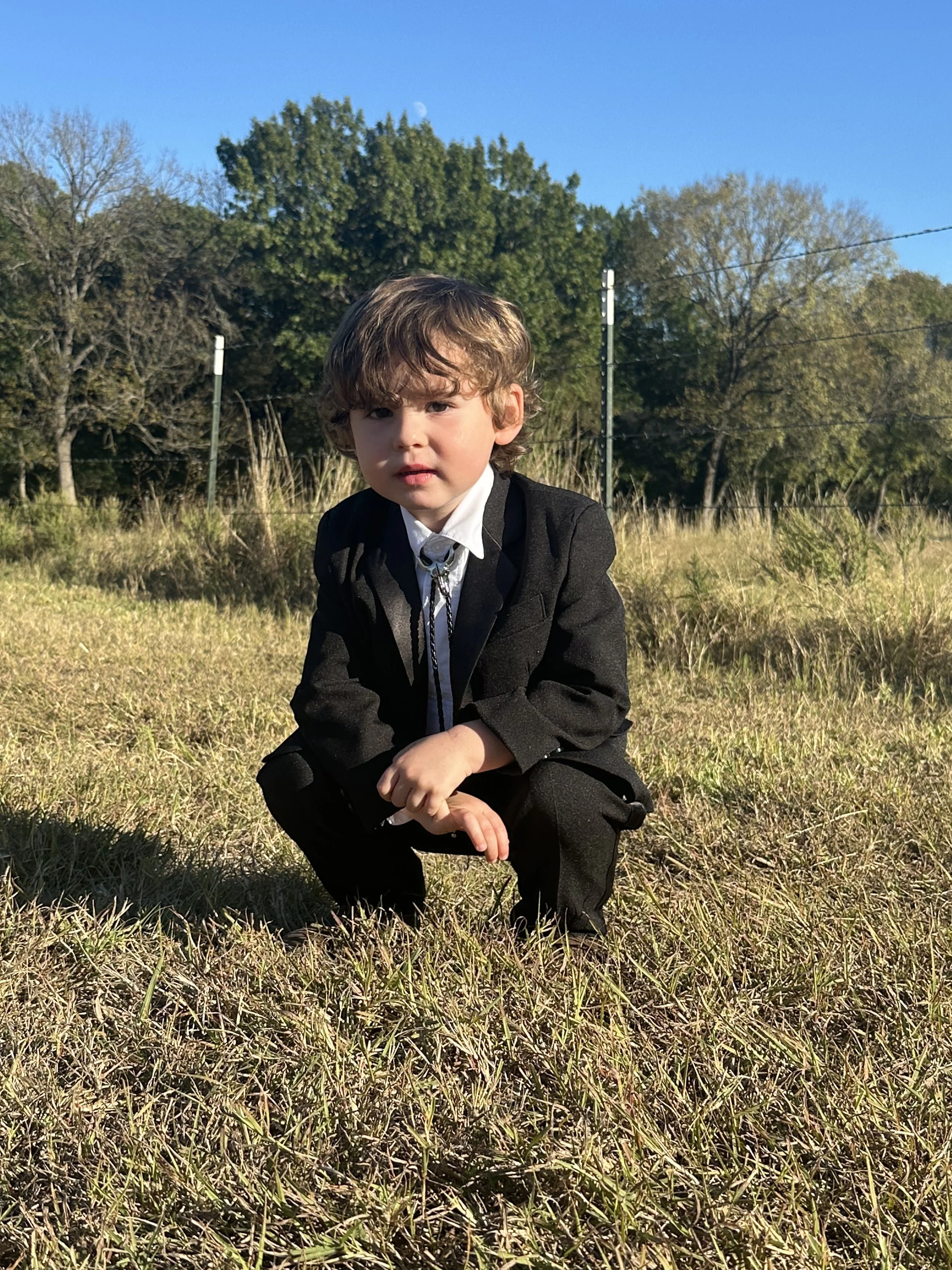A young boy crouching outdoors on a grassy field wearing a black suit and white shirt, with trees and a blue sky in the background.