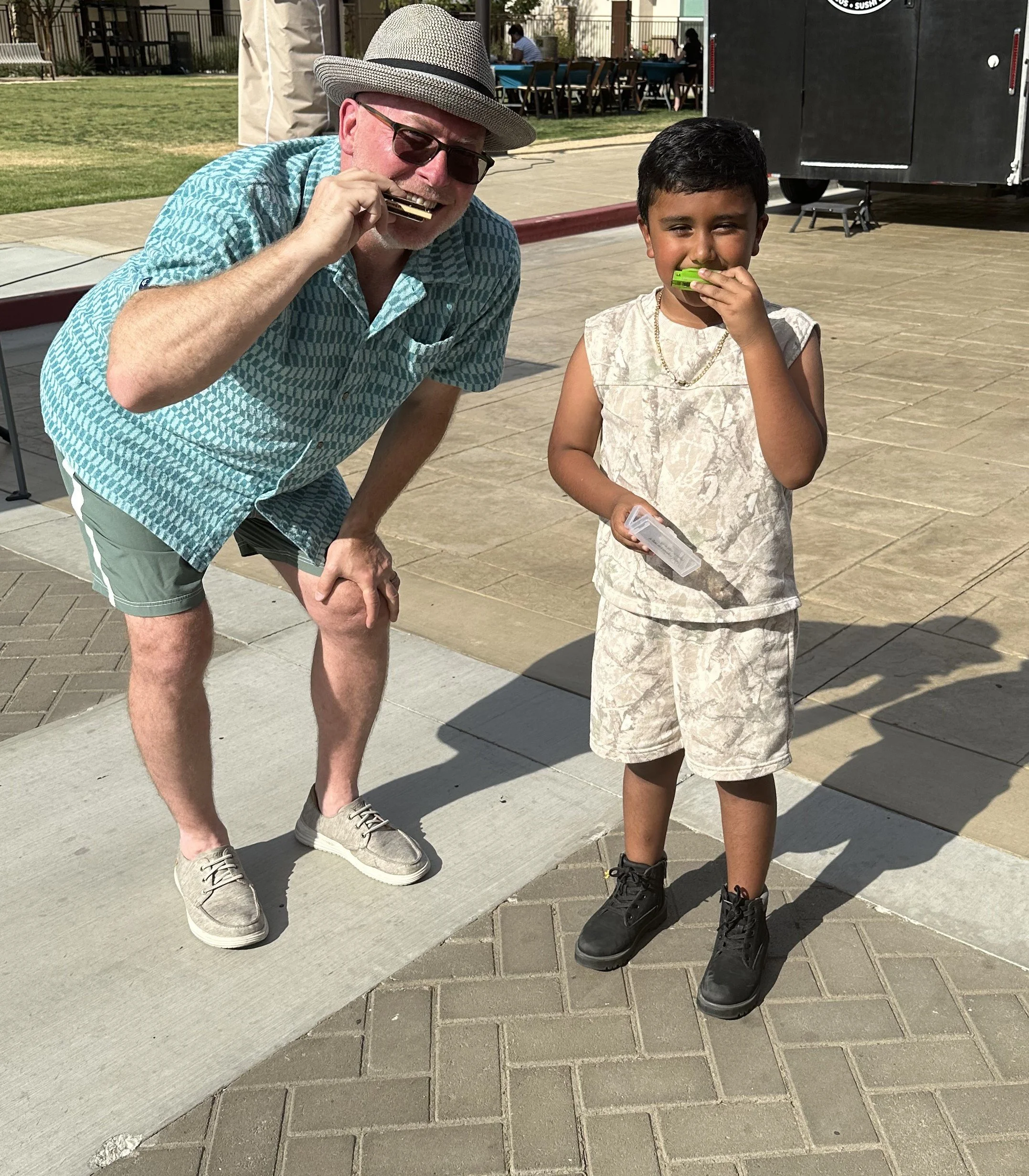 Two people, an older man and a young boy, are standing outdoors on a paved area. They are both holding and playing small harmonicas, smiling at the camera. The man is wearing a hat, sunglasses, a patterned short-sleeve shirt, and shorts. The boy is wearing a light-colored shirt and shorts, with black boots and a necklace.