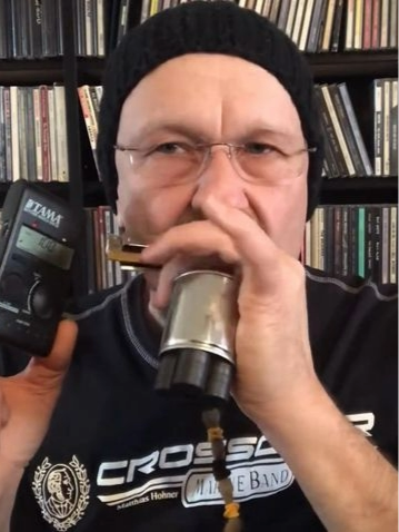 A man (Joe Filisko) wearing a black beanie and glasses, holding a microphone and a harmonica, standing in front of bookshelves.
