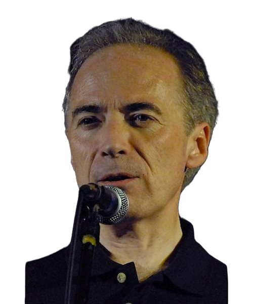 A man speaking into a microphone on stage, with a dark background and wearing a black shirt.