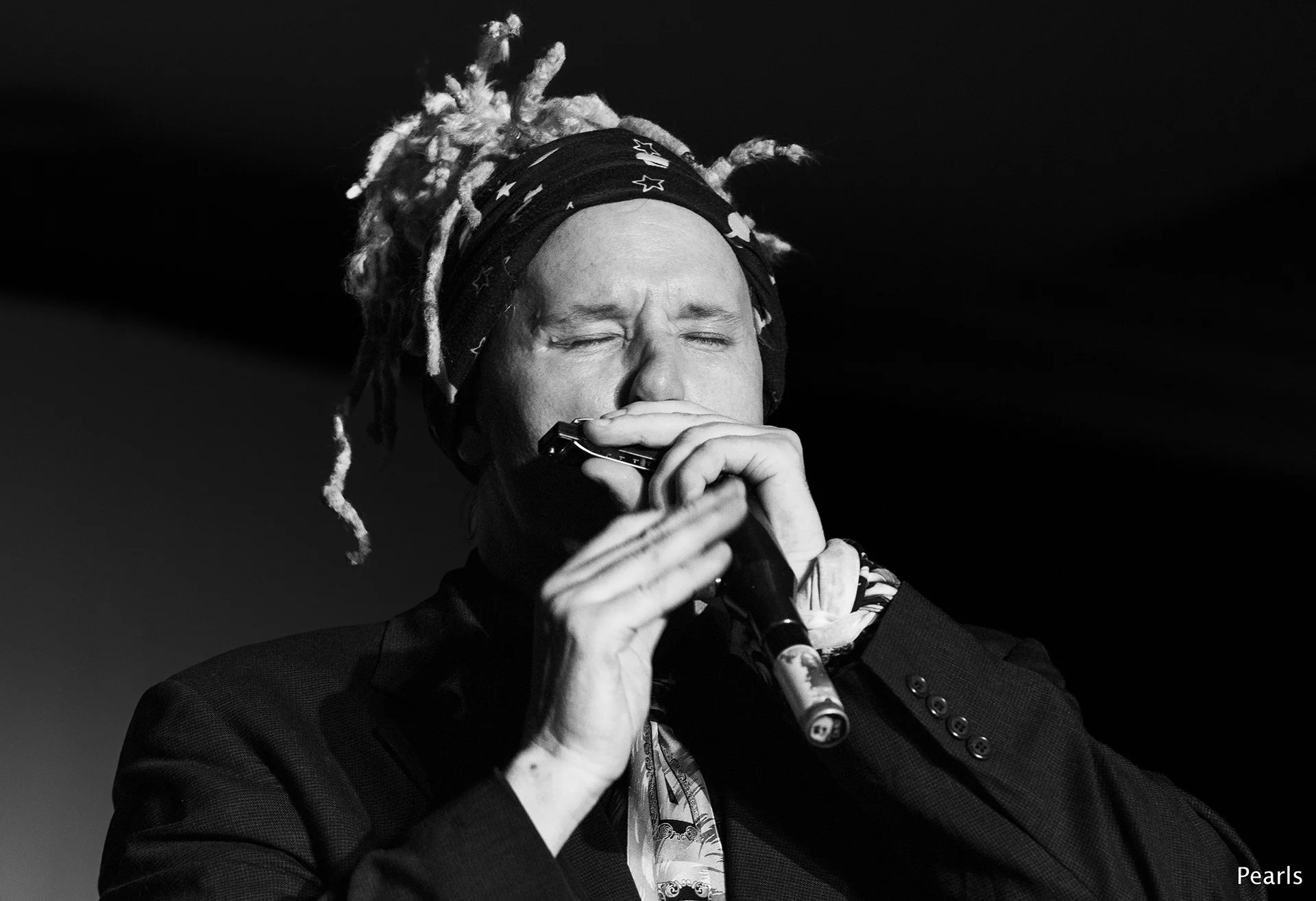 A black and white photo of a man, Jason Ricci, with light-colored dreadlocks and a bandana with stars. They are wearing a dark jacket and holding a harmonica near their mouth with eyes closed, playing music.