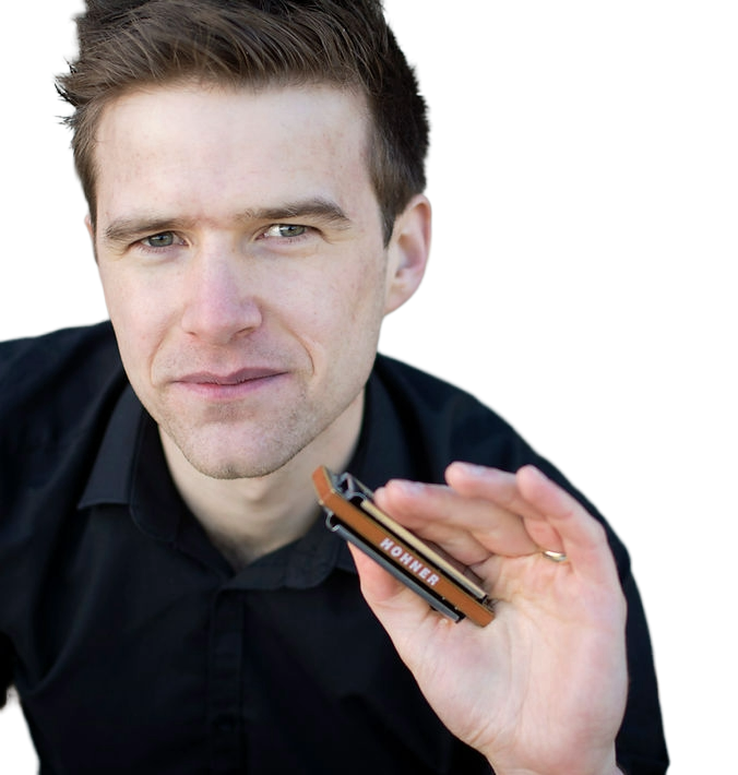 A man with short brown hair and light skin holding a harmonica up close to the camera, wearing a black shirt.
