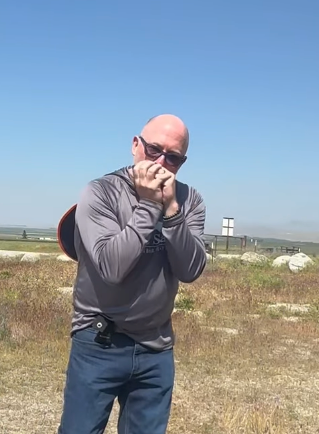 A man standing outdoors in a grassy area with a clear blue sky, wearing sunglasses, a gray sweatshirt, and jeans, holding his hands close to his face in a playful or shy pose.