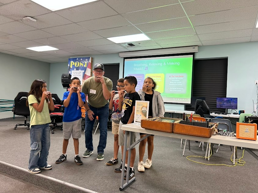 A group of children and an adult in a classroom or community room, engaging in a music activity. The children are standing, some with their hands near their mouths, and the adult appears to be leading or instructing. There is a large screen on the wall displaying instructions about breathing, relaxing, and making music, with colorful visuals.