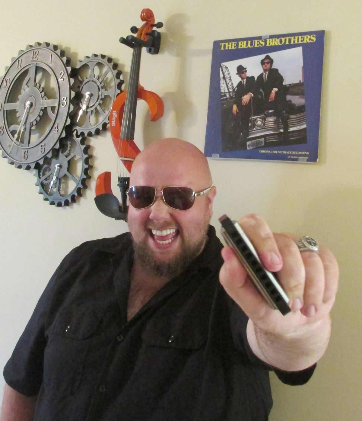 A man with a bald head, beard, and sunglasses smiling and pointing a remote control toward the camera. Behind him on the wall are clock gears, a toy orange violin, and a record album of The Blues Brothers with two men in black suits and sunglasses sitting on a car.