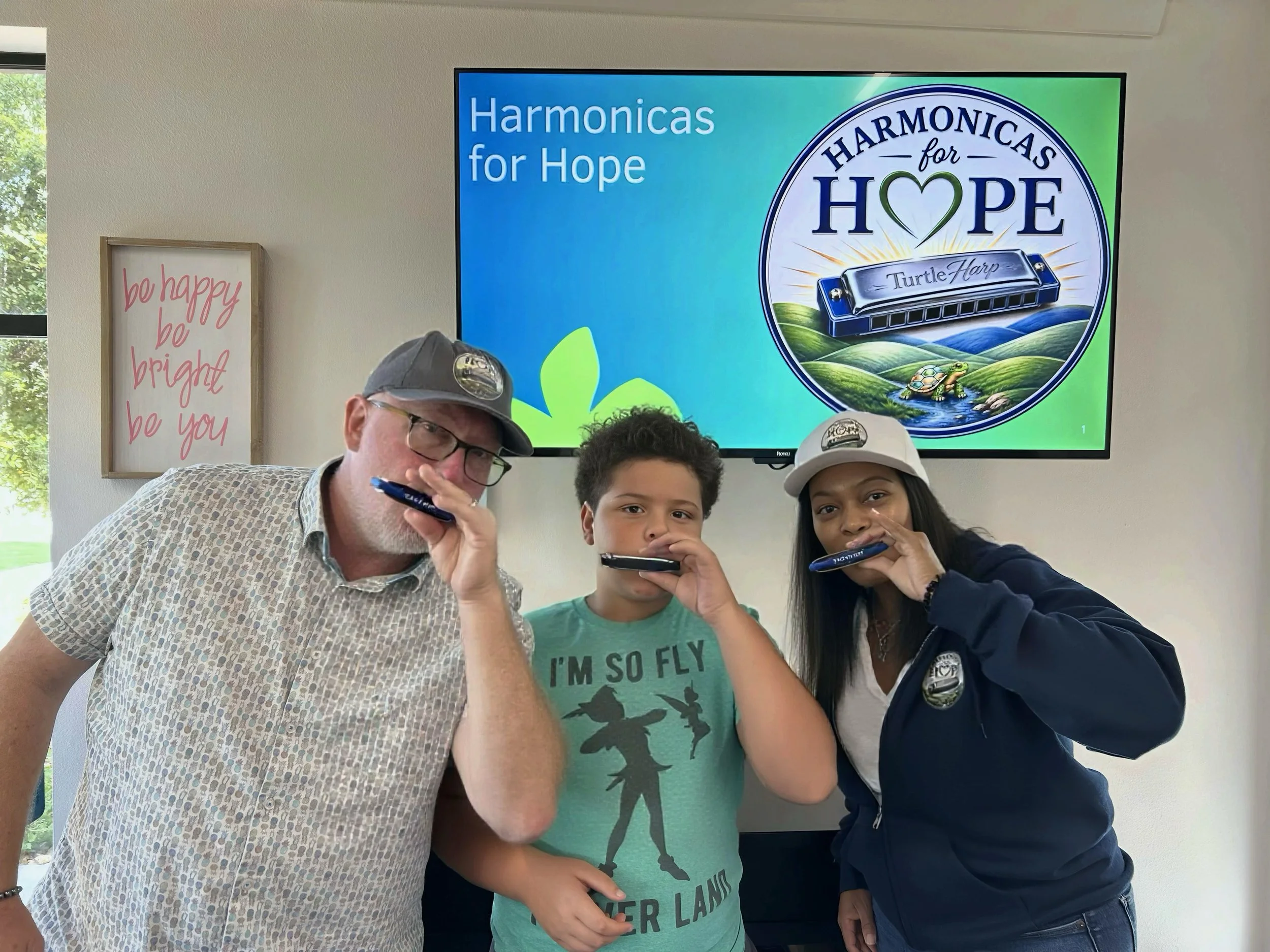 Three people, an older man and two children, pose with their faces close to the camera, holding markers in their hands. Behind them is a large TV screen displaying a logo for 'Harmonicas for Hope,' featuring a green heart in the word 'Hope,' and an illustration of a turtle-shaped harmonica set in a landscape of rolling green hills and a turtle. There is a window with tree branches outside to the left and a framed sign that says 'be happy be bright be you' on the wall.