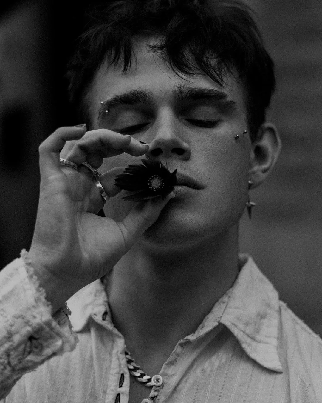 Portrait en noir et blanc d'un jeune homme avec des piercings, portant une chemise décontractée et des bijoux, tenant une fleur près de la bouche avec un regard fermé.