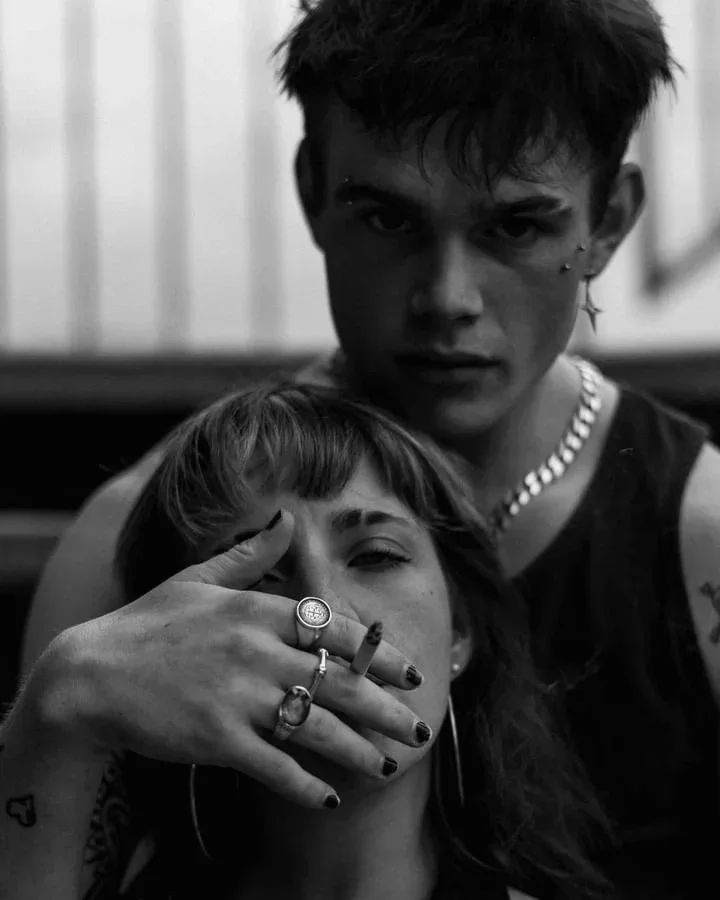 Un jeune homme et une jeune femme posent pour une photo en noir et blanc. Le jeune homme regarde directement l'objectif, tandis que la jeune femme cache en partie son visage avec sa main, portant plusieurs bijoux et des tatouages visibles.