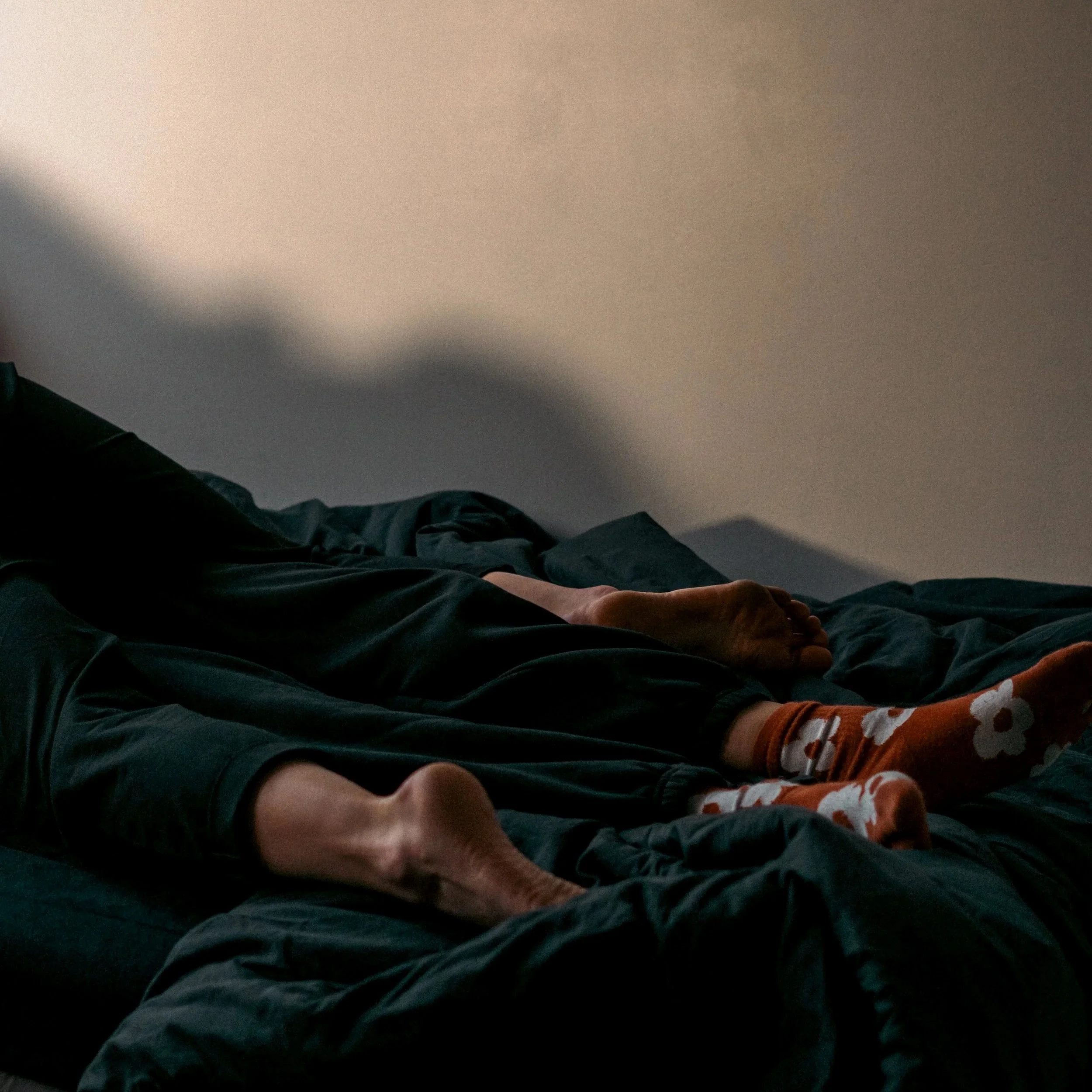 Une personne allongée sur un lit avec des vêtements sombres, portant des chaussettes à motifs blancs avec orange et noir, dans une pièce sombre avec un mur beige aléatoirement éclairé.