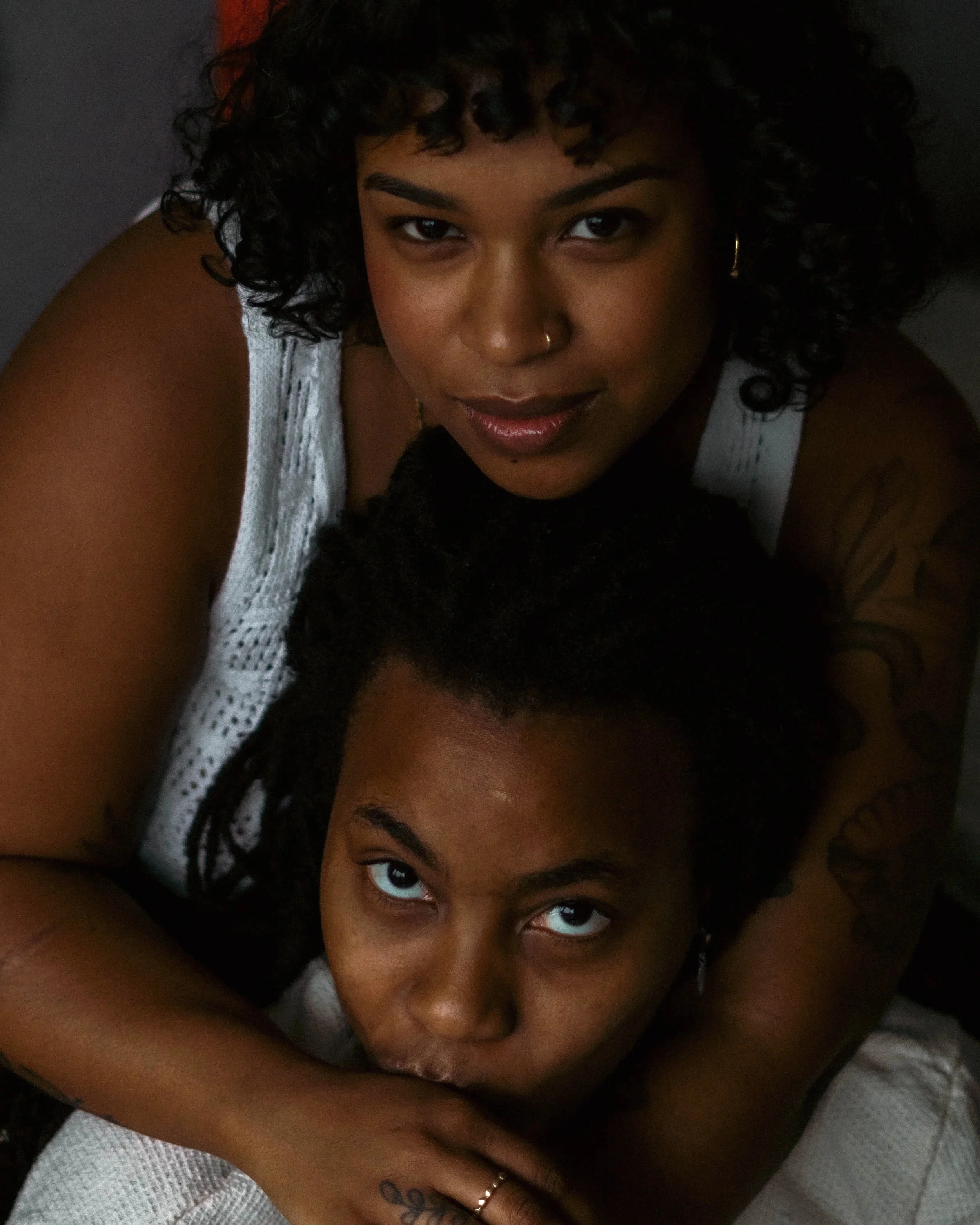 Gros plan d'une femme noire avec des cheveux bouclés, portant un maillot blanc, regardant droit devant avec un sourire subtil, dans un décor sombre.