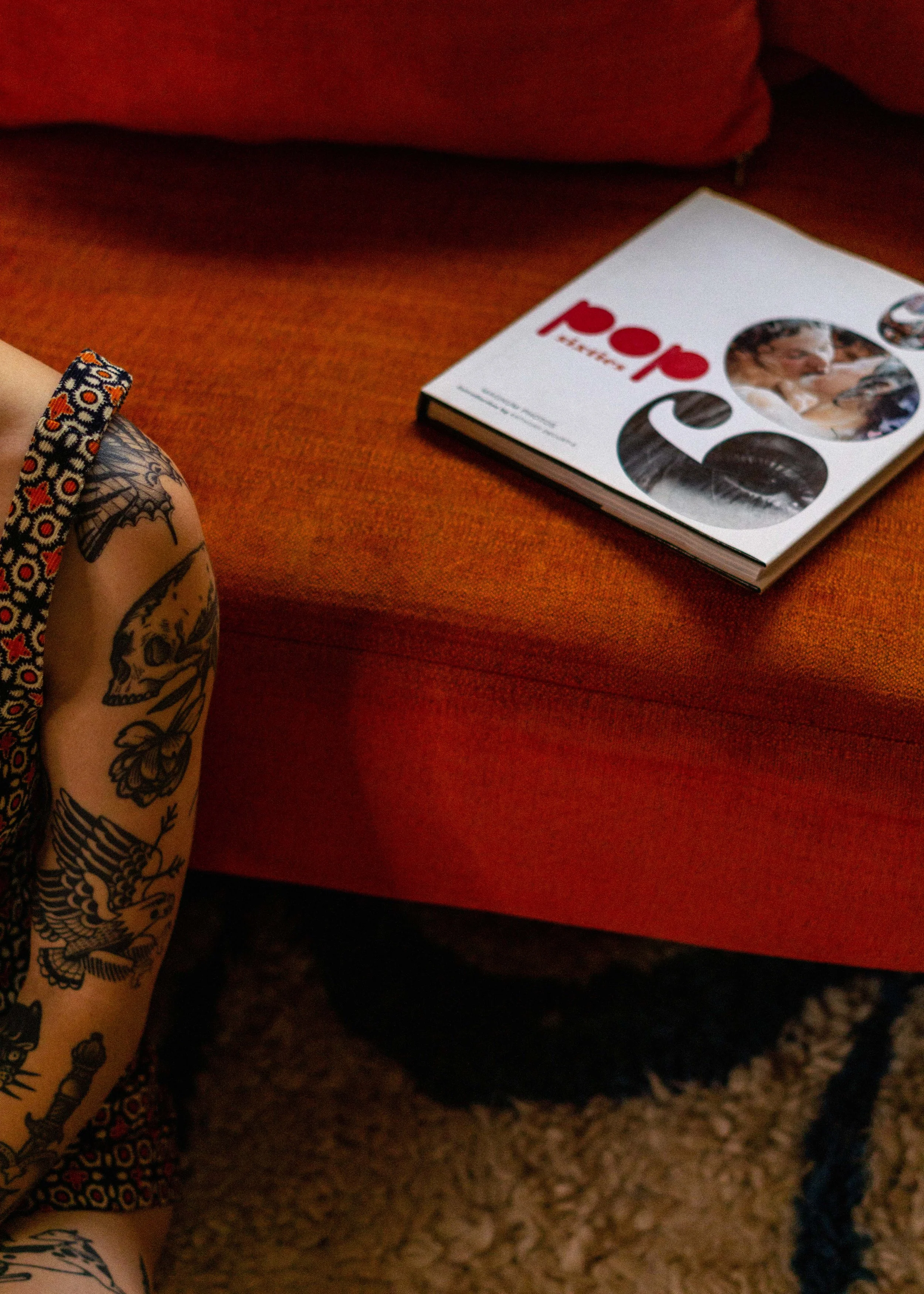 Une personne avec un bras tatoué portant un bracelet à motif floral, assise à côté d'une table rouge. Sur la table, il y a un magazine intitulé 'POP' avec des photos en couleuros et une femme souriante.