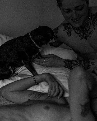 Une personne allongée dans un lit, une femme avec des tatouages joue avec un chien noir de petite taille. La scène est en noir et blanc.