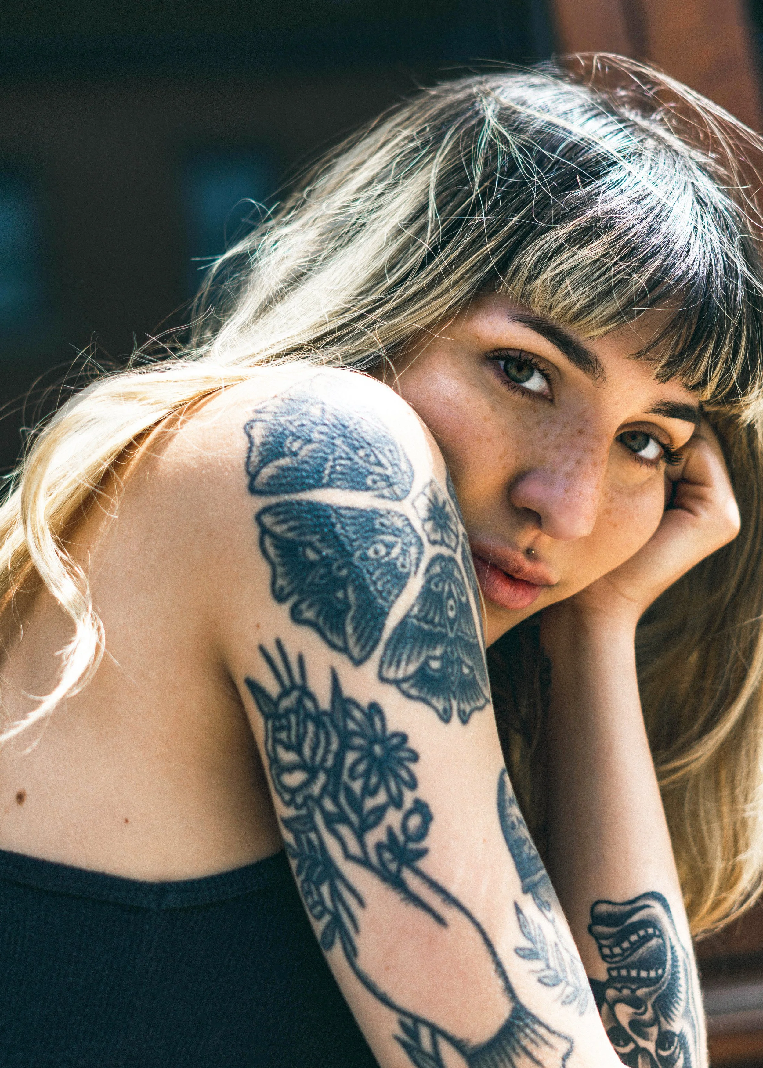 Une femme avec des cheveux longs blond et une frange, qui repose sa tête sur sa main. Elle a un tatouage de papillon noir sur son épaule et un tatouage de crâne sur son bras.