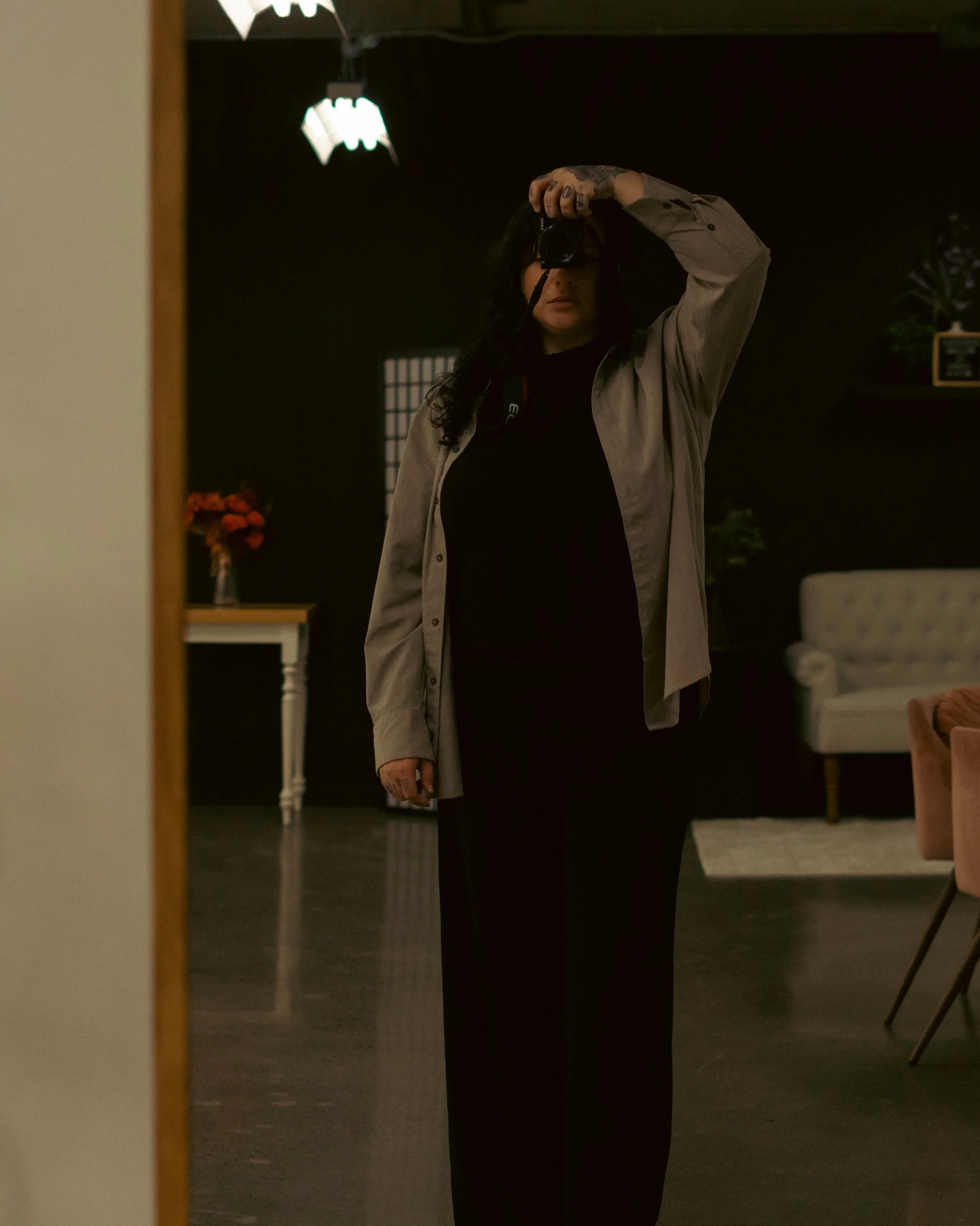 Un miroir reflétant une femme avec des cheveux noirs, portant une chemise beige, dans une pièce sombre avec des décorations et meubles.