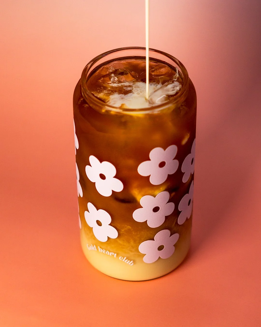Une canette de soda avec un motif de fleurs blanches sur un fond orange, avec du sirop ou une boisson claire vers le haut. La canette porte l'inscription 'wild heart club'.