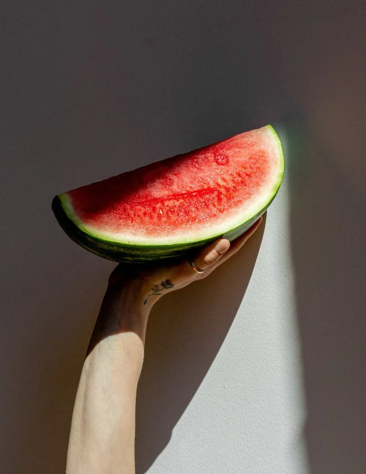 Une main tenant une grosse tranche de pastèque contre un mur blanc, avec des ombres nettes.