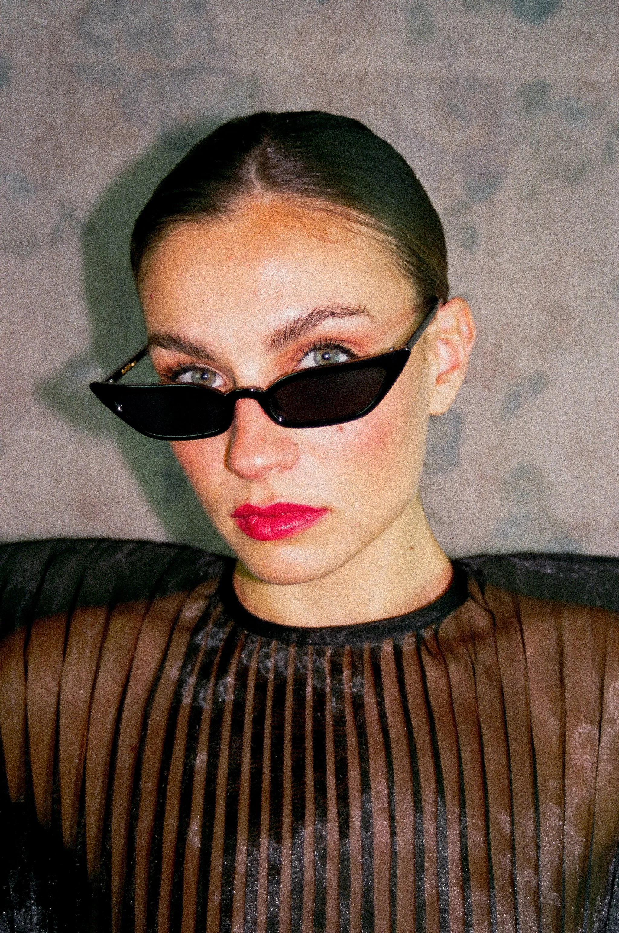 Une femme avec des lunettes de soleil noires, des cheveux tirés en arrière, portant un maquillage avec du rouge à lèvres rouge et un chemisier noir en filet avec des rayures verticales.